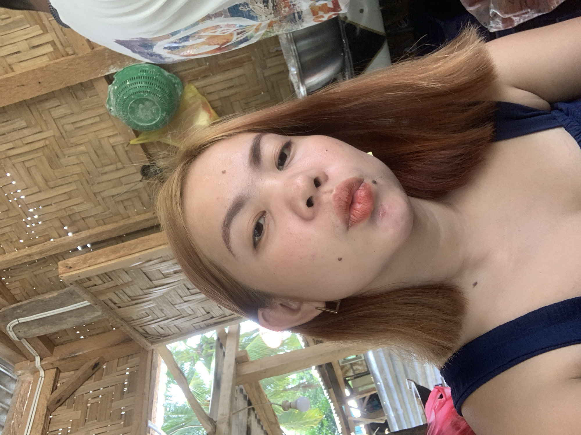 A young woman with shoulder-length auburn hair and fair skin is captured in a close-up selfie. She is wearing a dark blue tank top and a square-shaped gold earring on her left ear. Her lips are pursed, and she has several small moles on her face. The background appears to be a rustic, open-air structure with woven bamboo walls and wooden beams. In the background, to the right of the woman, a portion of another person's torso wearing a white t-shirt with a colorful graphic print is visible, along with a green, perforated plastic basket and a stainless steel bowl. To the left of the woman, palm trees and parts of a corrugated metal roof are visible, suggesting an outdoor or semi-outdoor tropical setting. The overall environment suggests a casual, possibly rural, location in Kiblawan, Philippines.