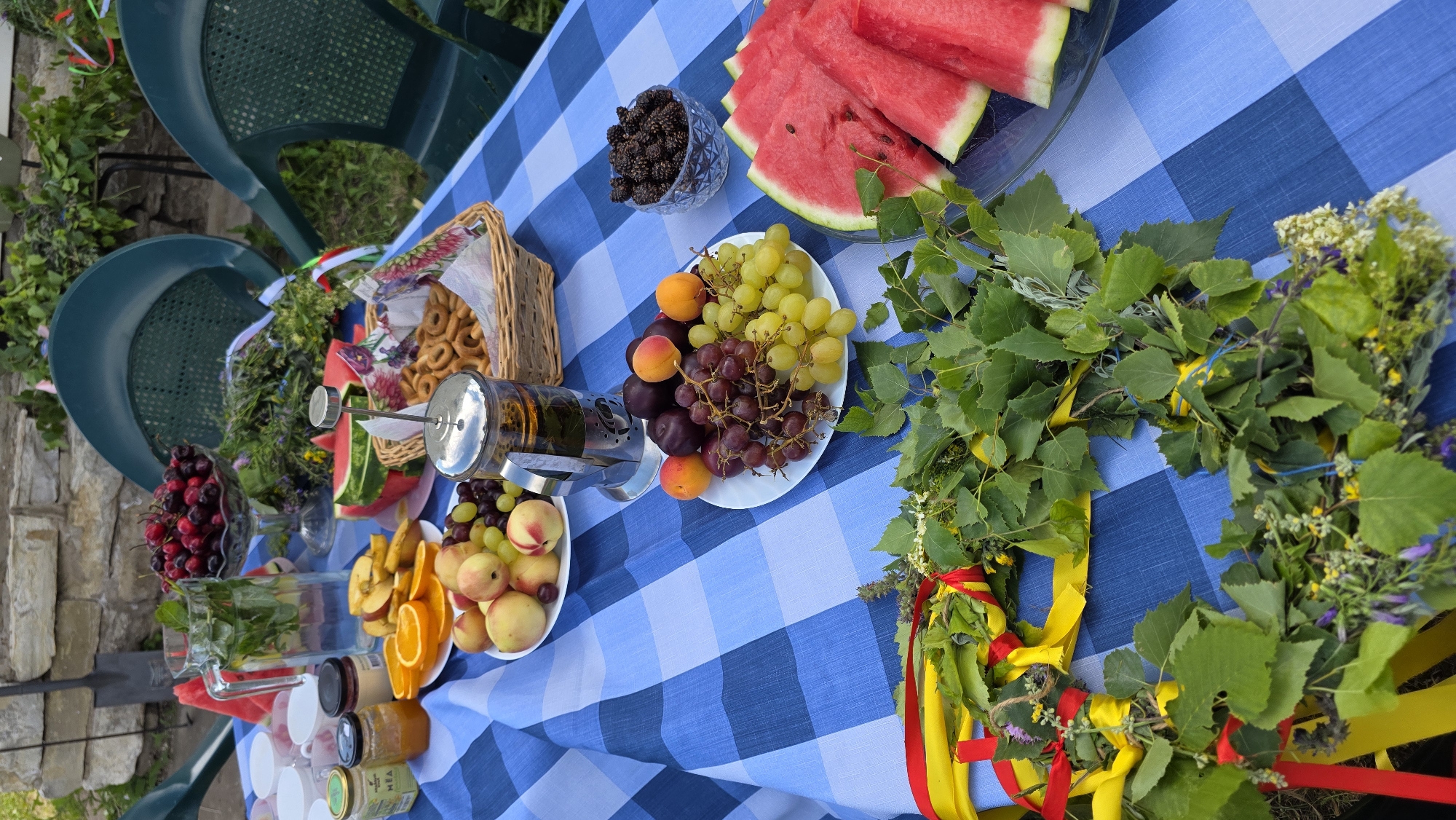 This image depicts an outdoor table setting in Kupoyarovo, Russia, laid with a blue and white checkered tablecloth. The table is laden with a variety of fruits, including sliced watermelon, peaches, plums, and bunches of green and red grapes. A glass pitcher filled with water and mint is also present, along with a glass bowl of cherries, a basket of small ring-shaped bread products, and a glass bowl filled with dark berries, possibly mulberries. A French press filled with tea or coffee sits on the table. In the foreground, a wreath made of greenery and adorned with red and yellow ribbons is laid out. The background shows a stone wall, green foliage, and two green plastic chairs. The overall scene suggests a gathering or picnic. No people are visible in the image.