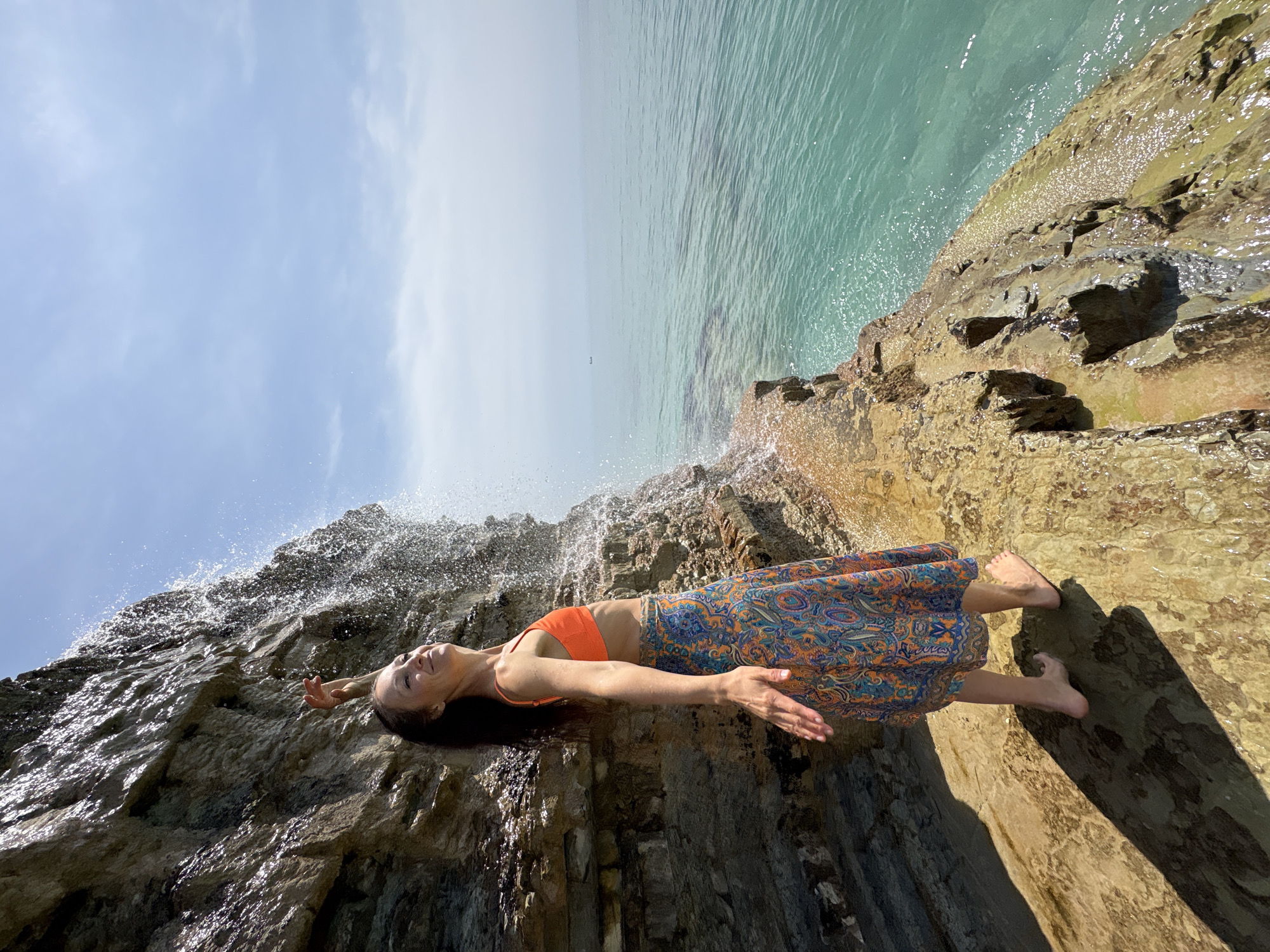 The image displays a woman lying supine on a rugged, wet rock formation under a bright sky. The scene is presented with a vertical horizon, placing the sky on the left, the rocky shore in the middle, and the sea on the right.
The central subject is a woman with dark hair, lying horizontally on her back. Her head is positioned towards the left, slightly elevated, and she has a peaceful, upward-gazing expression. She is dressed in an orange bikini top and a long, flowing skirt adorned with an intricate blue, orange, and yellow paisley-like pattern. Her arms are outstretched to her sides with open palms, and her bare feet are visible towards the right side of the frame, resting on the rocks.
Above and around the woman, water actively splashes and cascades over the dark, layered rocks, creating a dynamic effect. These rocks appear slick from the water and exhibit various shades of brown and grey, forming a textured, irregular surface. Adjacent to the rocky outcrop on the right, a calm, clear expanse of turquoise sea stretches towards a hazy horizon. The left side of the image reveals a bright, clear blue sky with a few wispy white clouds.
The strong sunlight indicates a daytime setting, likely midday or early afternoon. The overall atmosphere conveys a sense of serenity and connection with nature in a coastal environment, consistent with a location such as Gelendzhik, Russia. There is no visible text in the image.