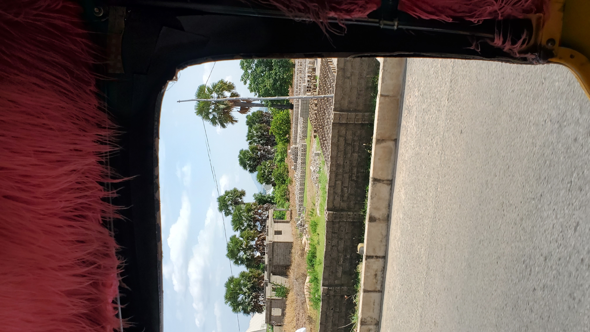 This is a photograph taken from inside a vehicle, possibly an auto-rickshaw, looking outwards. The foreground is dominated by the fuzzy, bright pink interior of the vehicle's side, obscuring the view to the left. The primary scene is viewed through a large opening on the right side of the vehicle.
The setting appears to be a semi-urban or rural area in Jalingo, Nigeria. The view reveals a landscape with several palm trees of varying heights. Behind the trees, there are partially constructed buildings, some with visible brickwork and concrete frames, suggesting ongoing construction. Piles of bricks are also visible, indicating a building material supply. Beyond the construction, there are more trees and a hint of distant hills or mountains under a bright, cloudy sky. A paved road runs alongside the scene on the right.
The time of day appears to be daytime, with bright sunlight casting shadows, though the exact time is not discernible. The weather seems fair with scattered clouds. There is no visible text in the image. The overall impression is one of observing a developing area on a clear day.
