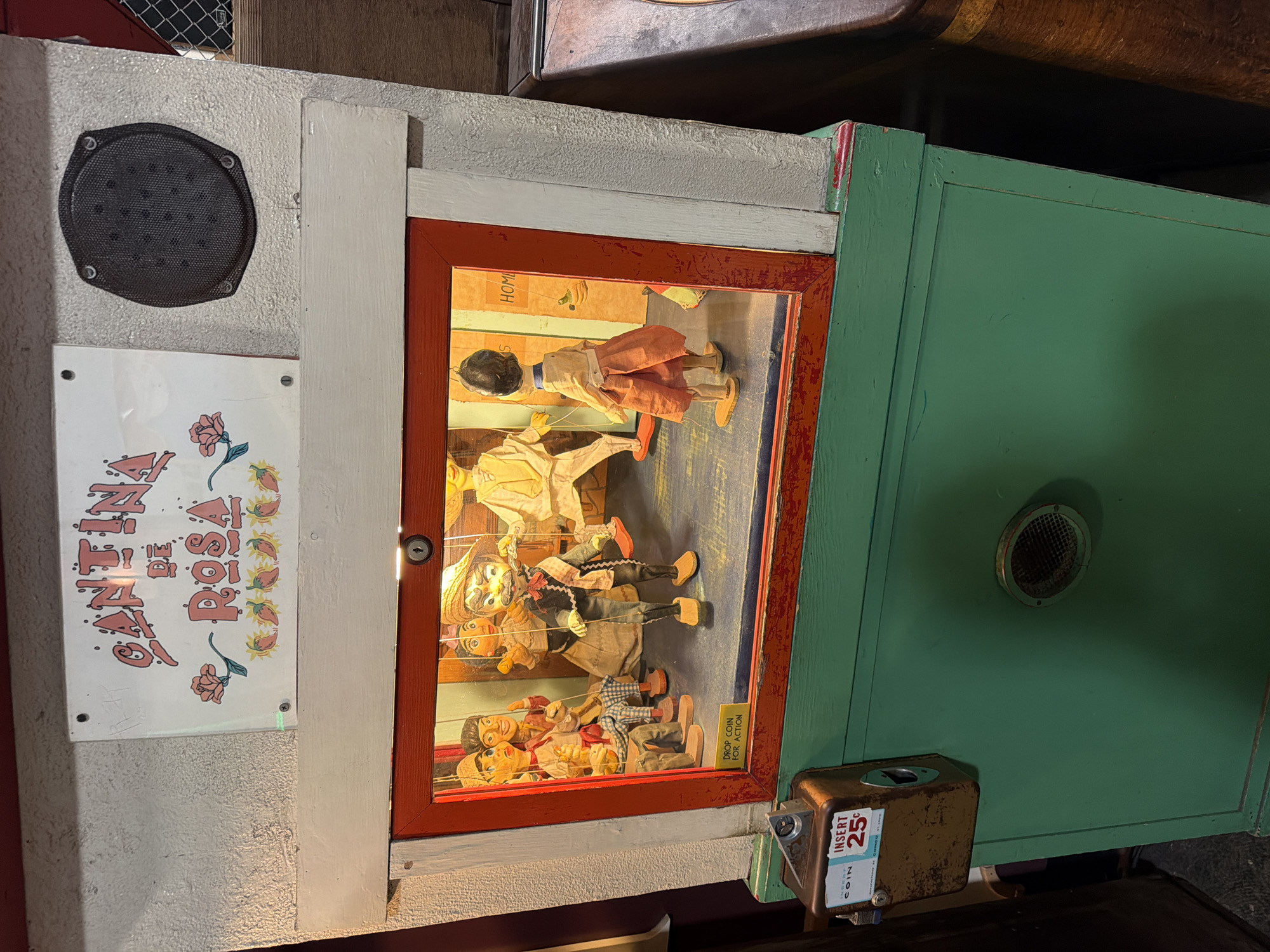 The image shows a vintage puppet theater with a sign above that reads "CANTINA DE ROSA". The theater is illuminated from within and displays several marionette puppets. The puppets are dressed in colorful clothing and are arranged in a scene that appears to be a performance. The theater is mounted on a green cabinet that has a coin slot labeled "INSERT 25¢". The overall aesthetic suggests an older, possibly handcrafted, attraction. The location is described as being in San Francisco, United States of America. The environment appears to be indoors, with the theater situated against a textured wall and next to a wooden cabinet.