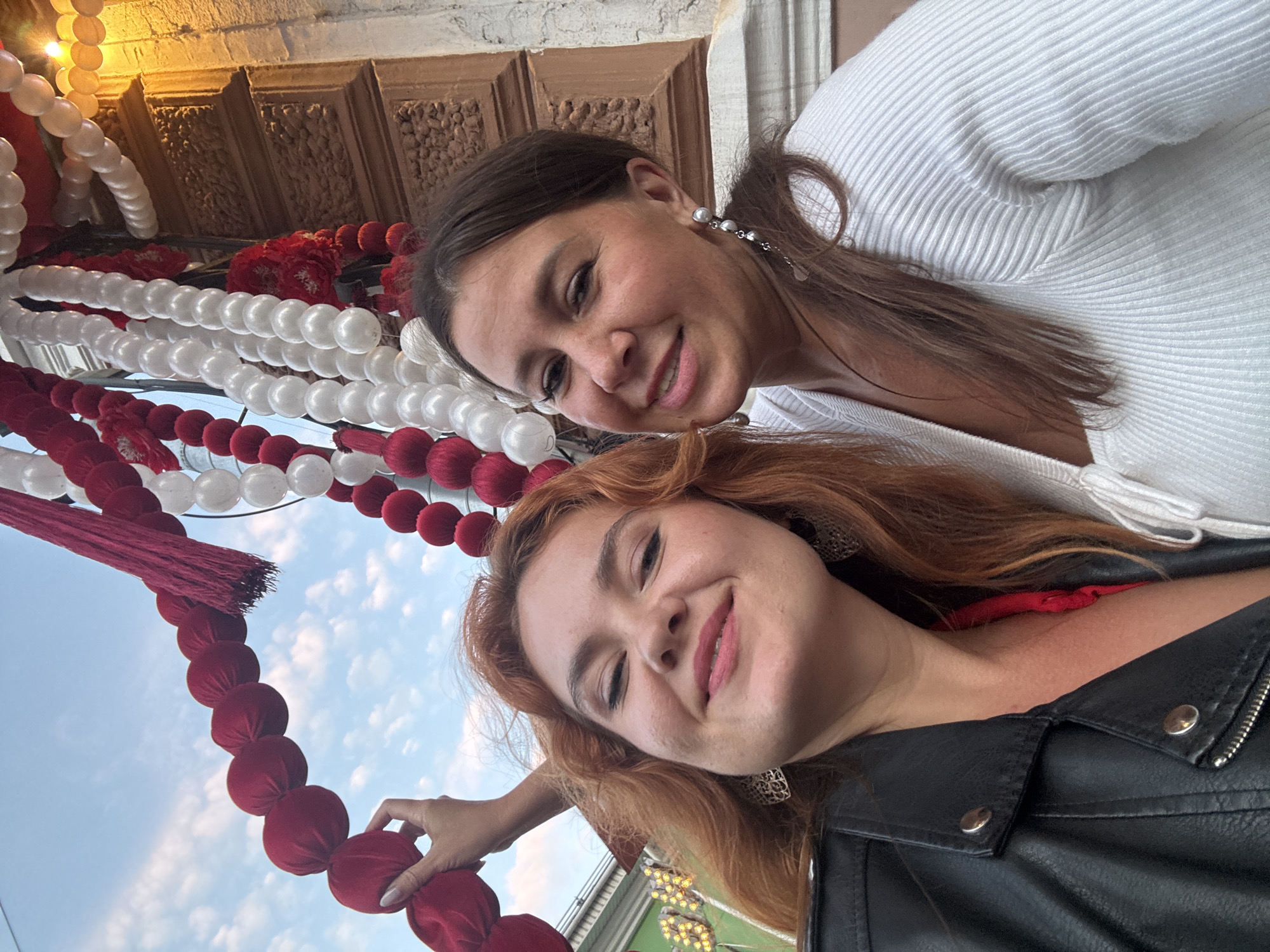 Two women are posing for a selfie outdoors in front of a building with ornate stonework and festive red and white decorations that appear to be balloon garlands. The woman on the left has auburn hair and is wearing a black leather jacket and a red top. She is smiling and has large, decorative earrings. The woman on the right has brown hair and is wearing a white ribbed top and pearl earrings. She is also smiling. The background includes a cloudy sky and a street lamp that is illuminated. The decorations suggest a celebration or event is taking place.