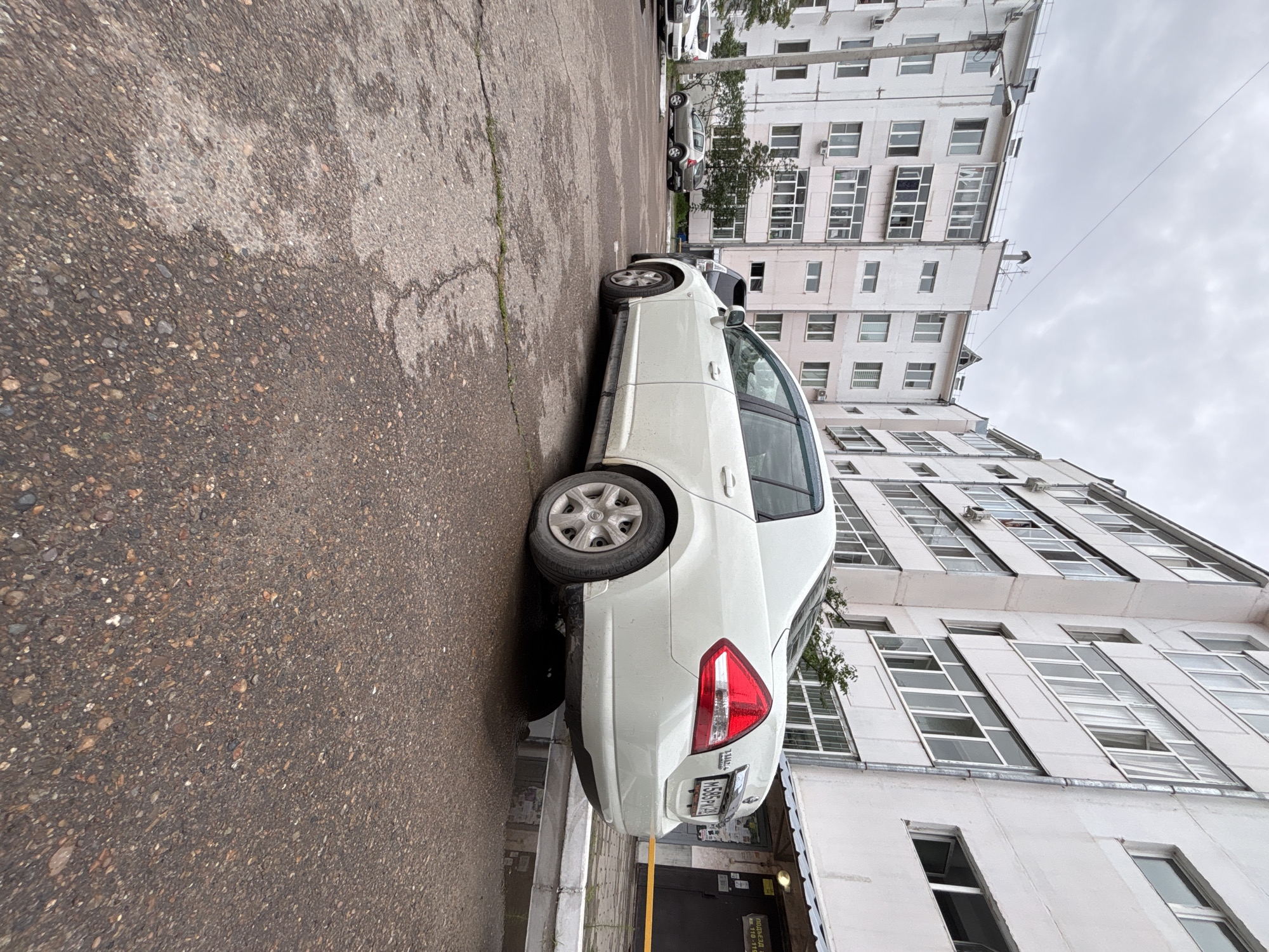 A white Nissan Tiida sedan is parked in a cracked asphalt courtyard. The car, with license plate M585PK, is seen from its rear quarter, facing right. The building behind the car is a multi-story apartment complex with white and pale pink facade, featuring numerous windows and balconies. In the distance, other cars are parked, and trees are visible. The sky is overcast, and puddles of water on the ground suggest recent rain. A yellow barrier tape is visible near the entrance of one of the building's sections.