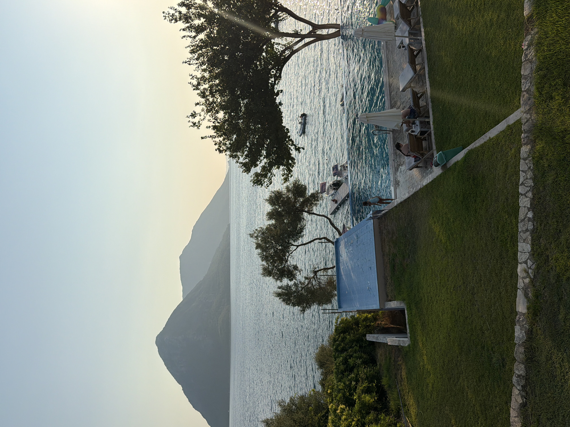The image captures a scenic view of a waterfront resort area in the Municipal Unit of Alyzia, Greece, during a clear, bright day with a pale blue sky. In the foreground, a well-maintained lawn slopes down towards a picturesque infinity pool that merges seamlessly with the calm, blue sea. The pool area is furnished with several lounge chairs, some occupied by people relaxing, and umbrellas providing shade.
To the left of the pool, a small, rectangular structure covered with a blue tarp is visible, possibly a smaller pool or a water feature. A wooden jetty extends from the shore into the sea, where a small motorboat is docked, and two other boats are anchored further out. The sea is a vibrant blue, with gentle ripples and the sun reflecting off its surface, creating a shimmering effect.
In the mid-ground, lush green trees and vegetation line the shore, with a particularly large, mature tree with dense foliage dominating the right side of the frame, its branches reaching out over the pool and sea. In the background, majestic mountains rise from the sea, their slopes covered in greenery, creating a dramatic and tranquil backdrop. The overall impression is one of a serene and idyllic vacation setting, bathed in the warm light of the sun.