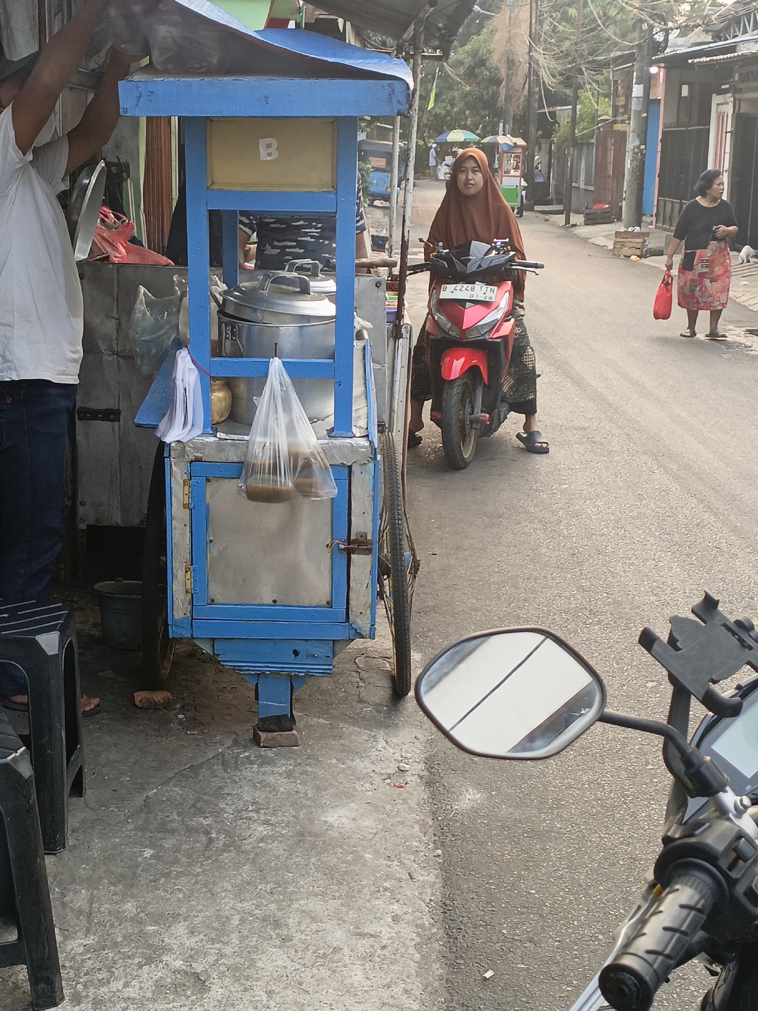 The image depicts a street scene in East Jakarta, Indonesia, during daylight hours. The foreground features a blue food cart with a yellow compartment labeled "B." A person, partially visible, wearing a white t-shirt and blue jeans, appears to be attending to the cart, possibly placing something into a plastic bag. Inside the cart, there are metal containers, likely for food preparation. Two plastic bags containing food are hanging from the cart.
In the middle ground, a woman wearing a brown hijab and a patterned skirt rides a red scooter. Her scooter has the license plate "B 4248 TJN." She is looking forward, appearing to move down the street.
In the background, the street is lined with buildings. Another woman, wearing a black top and a patterned skirt, is walking away from the camera, carrying a red plastic bag. The environment suggests a typical urban street in Indonesia with overhead wires and some vegetation visible. The overall lighting indicates it is likely late afternoon.