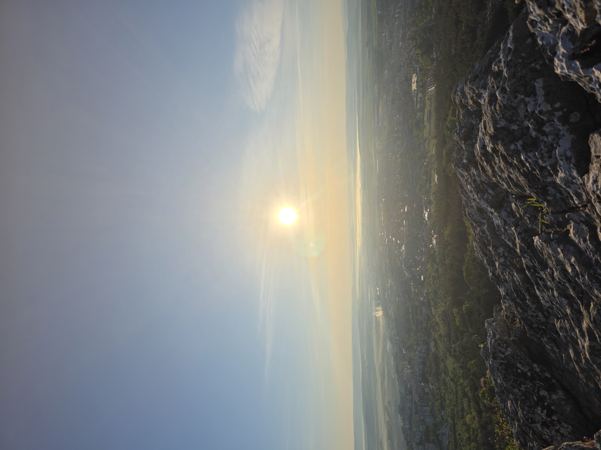 The image captures a view from a rocky elevation, possibly a cliff or mountainside, overlooking an urban or suburban area shrouded in morning mist. The sun is positioned high in the sky, emanating a bright glow that partially obscures the landscape below. The city is characterized by a mix of residential buildings, greenery, and possibly industrial structures, though the mist obscures details. The foreground is dominated by jagged rock formations, with sparse vegetation clinging to the cliffside. The perspective suggests the image was taken from a higher vantage point looking down towards the city.