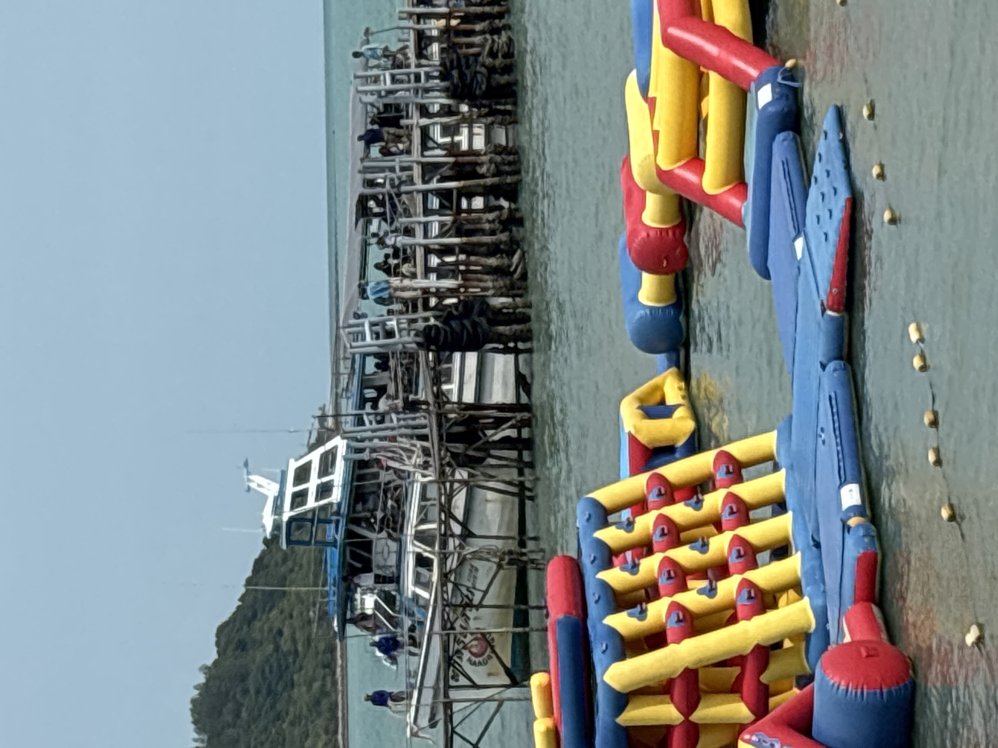The scene depicts a waterfront in Baan Bang Rak, Thailand. A pier constructed of weathered wood and metal pilings supports a multi-level structure, possibly a fishing platform or a docking area. A white and blue boat is moored alongside the pier. Several people are visible on the pier structure. In the foreground, an inflatable water park structure with red, yellow, and blue elements floats on the water's surface. The water appears calm. A green, hilly landmass is visible in the background, under a clear, light blue sky.