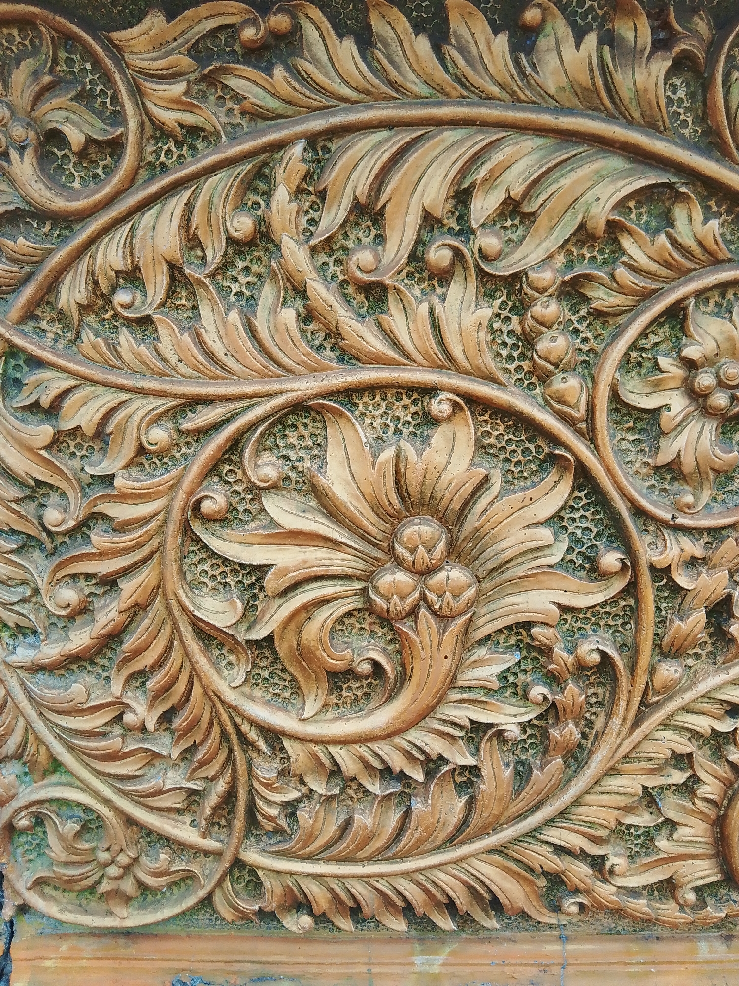 The image displays a close-up, detailed view of a decorative carved panel, likely made of wood or a similar material, with a golden-brown finish. The carving features an intricate floral and foliate motif, characteristic of Javanese art. Elaborate scrollwork intertwines with stylized leaves and small clusters of berries or buds. The background of the carving is textured with a fine, honeycomb-like pattern, adding depth and contrast to the raised elements. The lighting accentuates the three-dimensional nature of the carving, casting shadows that highlight the contours and details of the design.
This style of carving is commonly found in the Tahunan district of Jepara Regency, Central Java, Indonesia, a region renowned for its high-quality wood carving traditions. These decorative panels are often used as architectural elements in traditional Javanese homes, temples, and public buildings, or as standalone art pieces. The specific design suggests a traditional or classical Javanese aesthetic, often associated with royal courts or significant cultural sites.