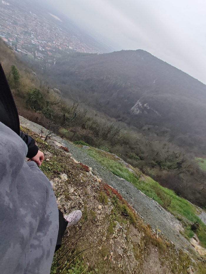 A high-angle photograph captures a scene from a cliff edge overlooking a valley and the city of Shumen, Bulgaria, under an overcast sky. The foreground features the lower body of an individual, dressed in grey sweatpants and a black jacket sleeve, with a light-colored sneaker visible on their right foot. Their right hand rests on their thigh. The individual stands on a steep, rocky incline covered with patches of green moss and reddish-brown vegetation. Immediately beyond, the terrain descends sharply into a valley densely covered with deciduous trees, largely bare, suggesting a winter or early spring season. In the midground, the valley floor gives way to a sprawling urban area identified as Shumen, characterized by numerous buildings and streets, fading into the atmospheric haze. On the far side of the valley, a forested mountain range rises, also blanketed in bare trees and obscured by the prevailing mist. The sky is uniformly grey and overcast, contributing to the muted color palette of the scene.