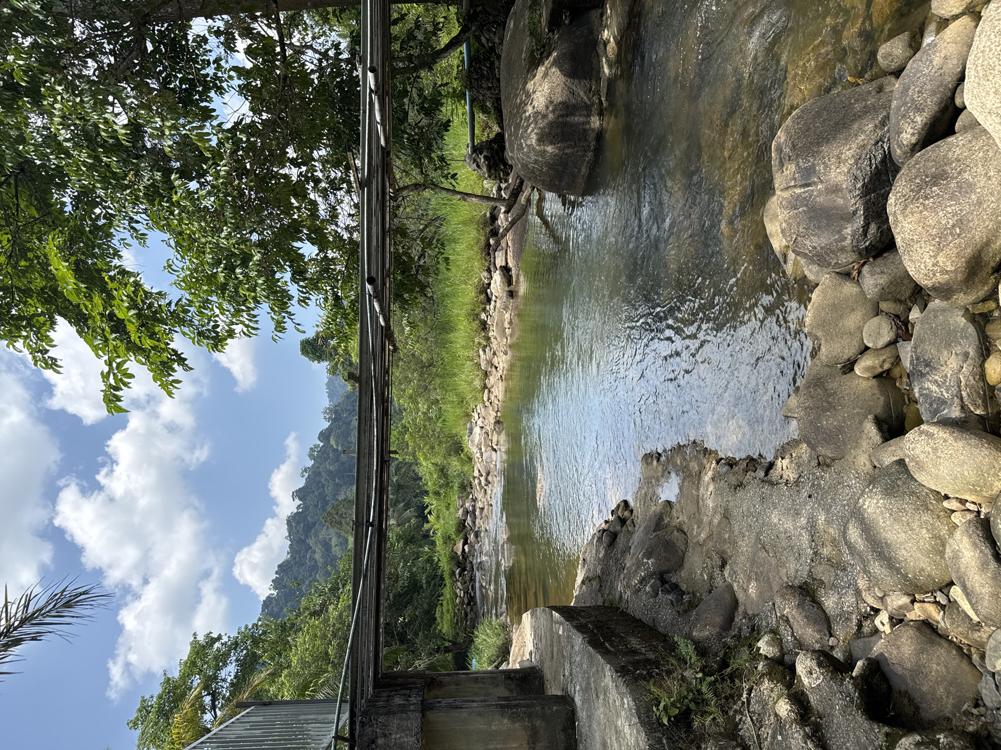 The image depicts a flowing river in a natural setting, likely taken in Tambon Song Phraek, Thailand. The river is bordered by rocks and vegetation. There is a man-made structure, possibly a bridge or support beam, spanning across the river. The sky is visible with scattered clouds. A mountain range is present in the background.