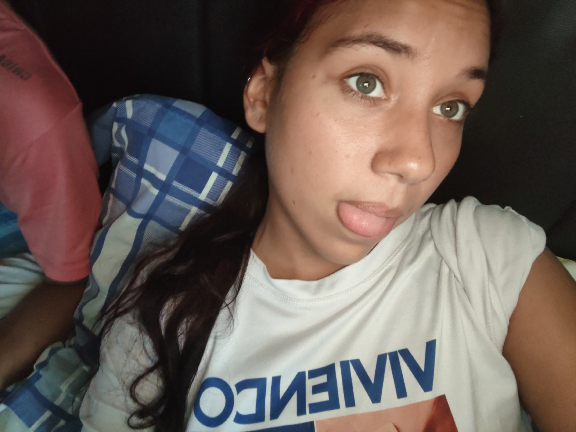 The image shows a young woman with brown hair and green eyes, sticking her tongue out. She is wearing a white t-shirt with blue lettering that appears to be reversed or a logo. She is lying down, possibly in bed, with a blue and white plaid blanket and a pink garment visible behind her. The background is dark, suggesting an indoor setting at night or in a dimly lit room. There is no visible indication of the location being Piloncito, Venezuela.