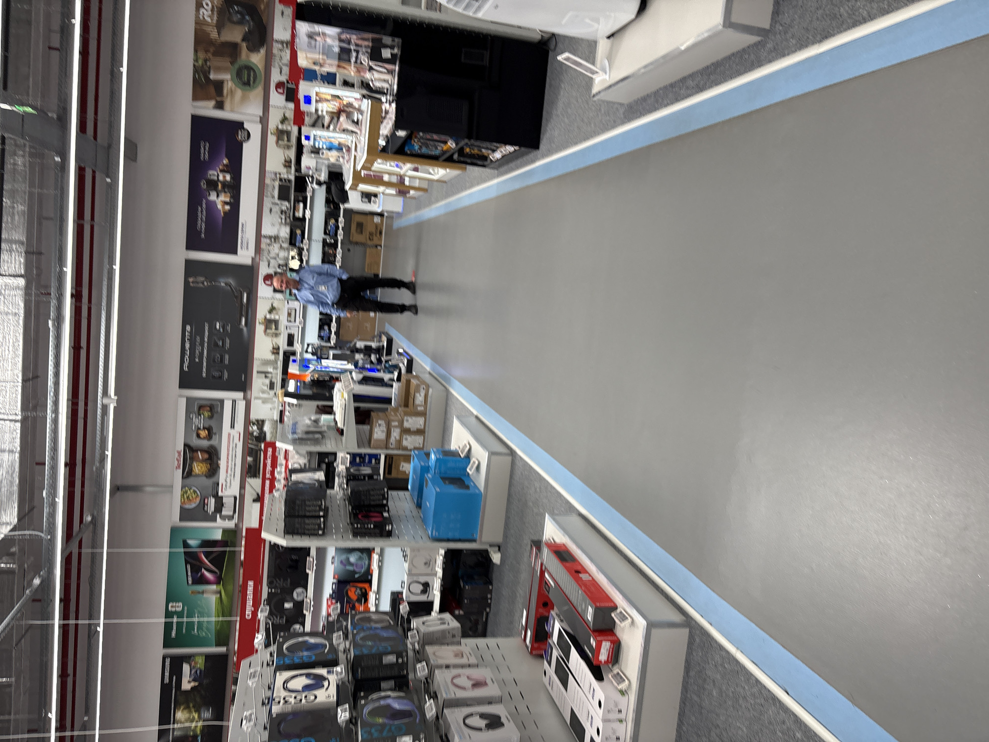 The image shows the interior of a large retail store, likely located in Lovech, Bulgaria given the request for location specificity. Fluorescent lights illuminate the aisles. The floor is grey with a blue accent line. Aisles are stocked with various electronics and household appliances. A man wearing a light blue shirt is walking down the aisle. Shelves are filled with products including headphones, kitchen appliances, and other consumer electronics. Store advertisements and product information are visible on the shelving. The scene appears to be in a well-organized retail setting.