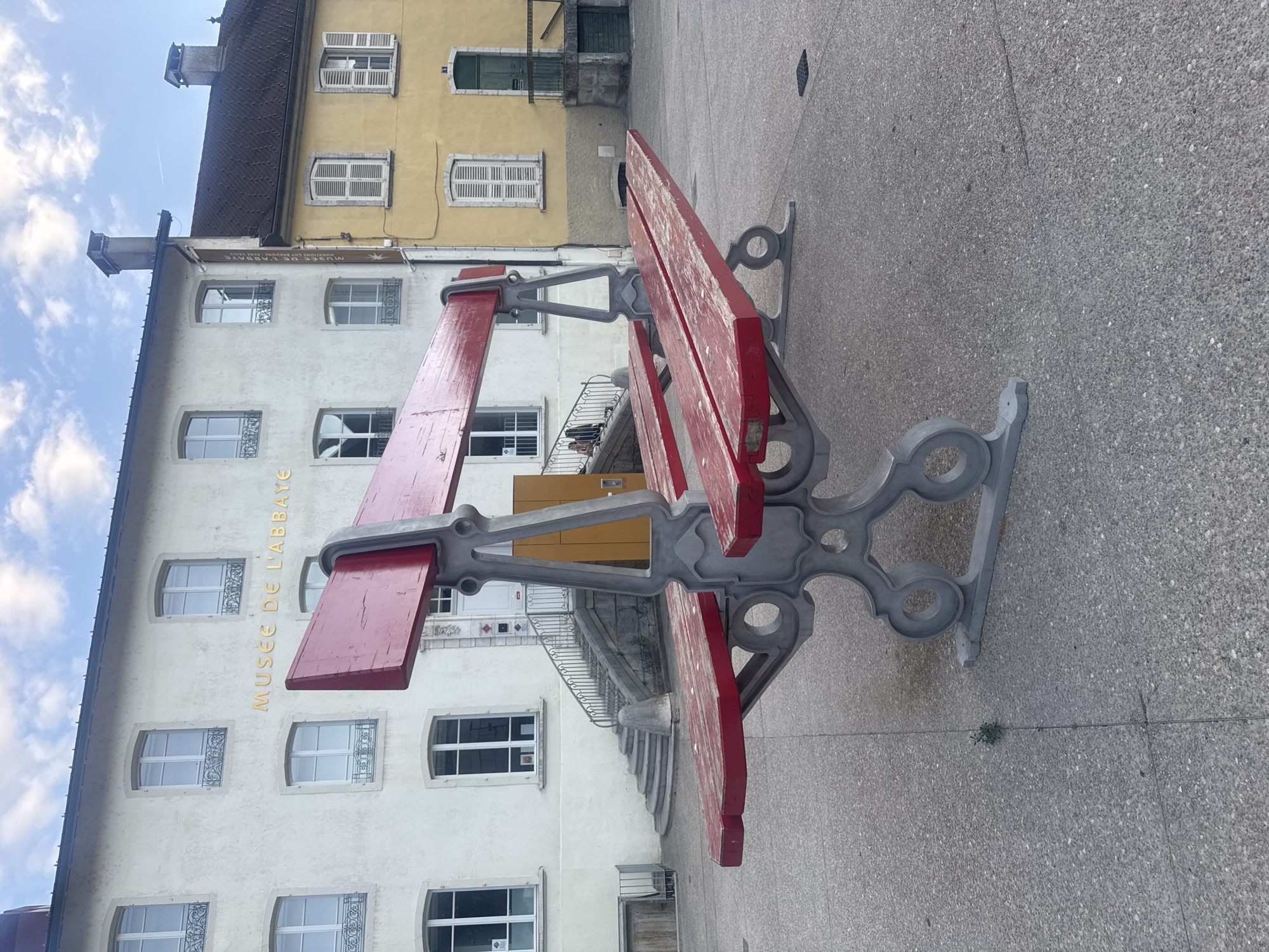 The image shows a large, oversized red bench with grey ornate metal legs in an outdoor plaza. The bench is the main subject and appears to be a public art installation. Behind the bench, there is a light-colored building with the words "MUSÉE DE L'ABBAYE" visible in gold lettering. This building has several windows with decorative ironwork. To the right of the light-colored building is a yellow building with a dark-tiled roof and shutters on its windows. The ground is paved with grey flagstones. In the background, a set of stone stairs leads up to the light-colored building. Two people are sitting on the stairs. The sky is partly cloudy. The scene is located in Saint-Claude, France, as indicated by the museum's name which is an abbey museum in Saint-Claude.