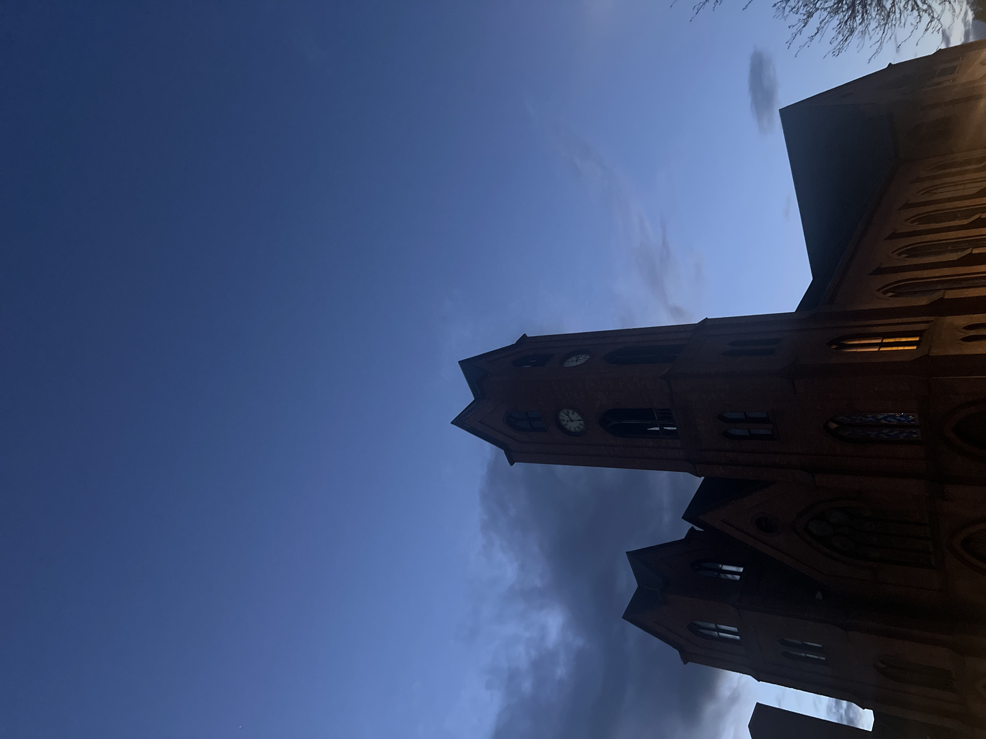 The image shows a brick church building against a blue sky. The church has a tall tower with clock faces visible on its sides. There are arched windows and other architectural details on the building. Some windows are illuminated from within. The sky is a gradient of blue, with some clouds present. The photograph appears to be taken from a low angle, looking up at the church. A few bare tree branches are visible in the upper right corner. The church is the Dutch Reformed Church, located in the city of Newburgh, New York.