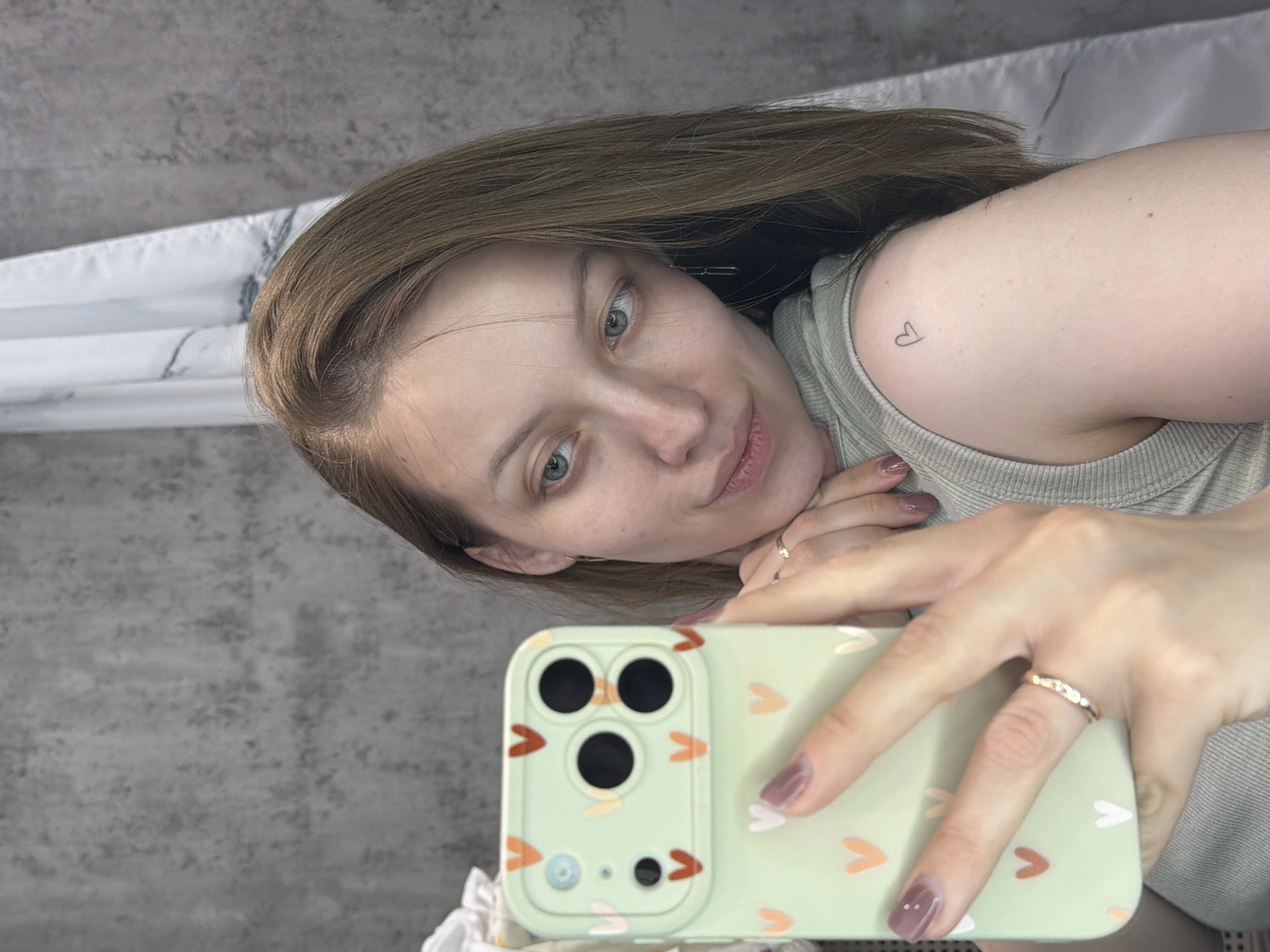 A light-skinned woman is holding up a green iPhone with a heart pattern case. The woman has shoulder-length blonde-brown hair. She is wearing a light-colored sleeveless shirt. There is a heart tattoo on her upper arm. She is wearing two rings. The background appears to be a grey textured wall. The location in the city of Reshetnikovo, Russia is unknown.