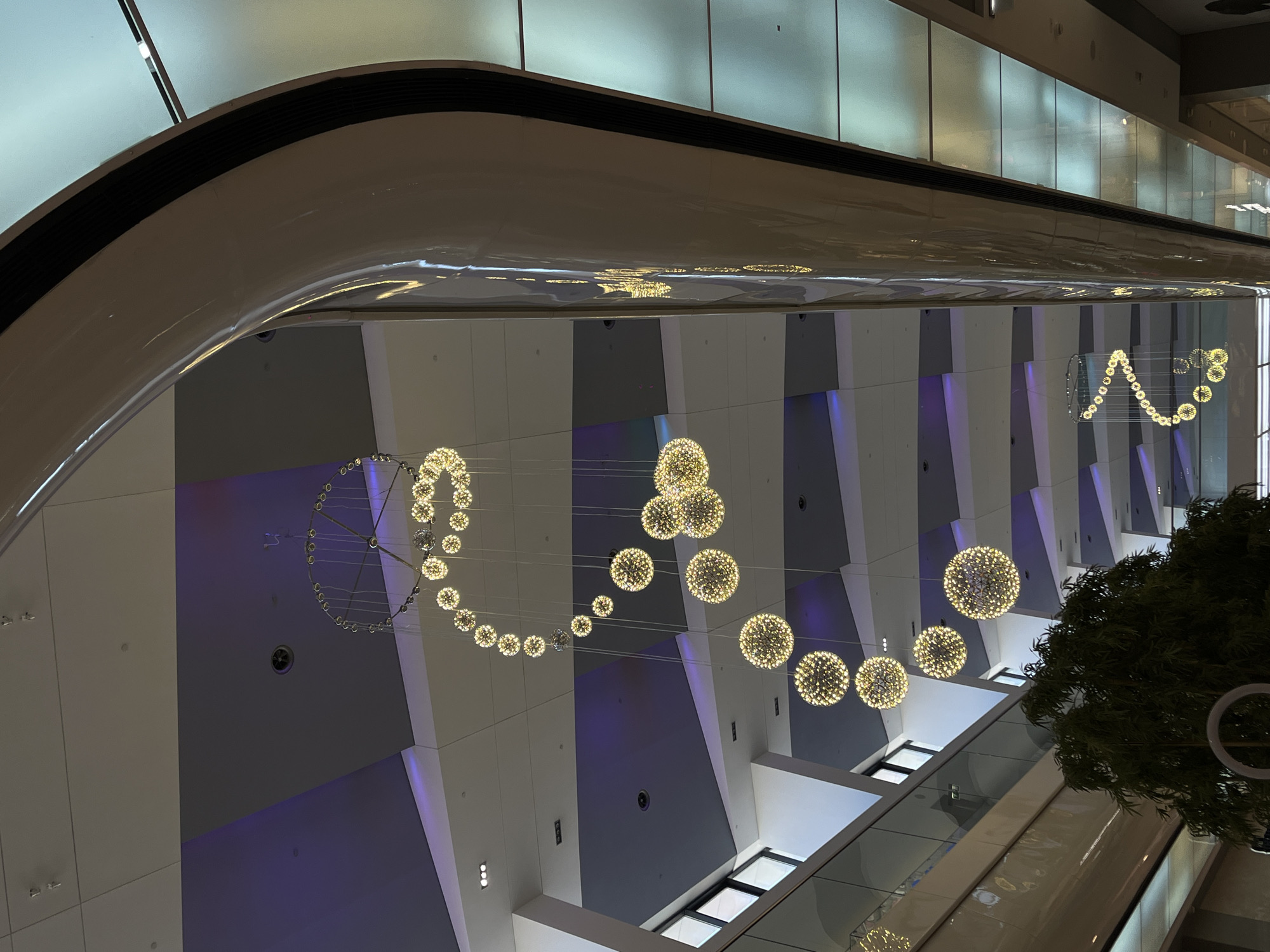 This upward-looking shot captures the modern interior of a building, likely a shopping mall or a large public atrium, located in Krasnodar, Russia. The scene is dominated by intricate, illuminated spherical light fixtures suspended from a high ceiling.
The main subjects are the decorative light installations, which appear as clusters of small, bright lights forming large, glowing spheres. Several of these spheres are arranged in a dynamic, undulating pattern across the vast ceiling space. In the foreground and extending to the left, a different style of lighting features smaller, individual light elements arranged in a circular or spiral pattern, connected by thin wires. Reflections of these lights shimmer on the sleek, curved white ceiling structure that frames the upper portion of the image.
Architecturally, the building features multiple levels with glass railings, creating an open and expansive vertical space. The walls on the upper levels are composed of large panels in varying shades of gray and white, some of which are accented with strips of subtle purple backlighting, adding a contemporary touch to the ambiance. A large, dark green plant or tree is partially visible on the right side, extending upwards, providing a natural element within the modern environment. Recessed rectangular lights and ventilation grilles are also visible on the ceiling and walls.
There is no discernible activity or people in the image; the focus is solely on the architectural design and lighting. The artificial illumination suggests an indoor setting where the time of day is not explicitly revealed, but the bright and sophisticated lighting creates a vibrant atmosphere. No text is visible in the image.