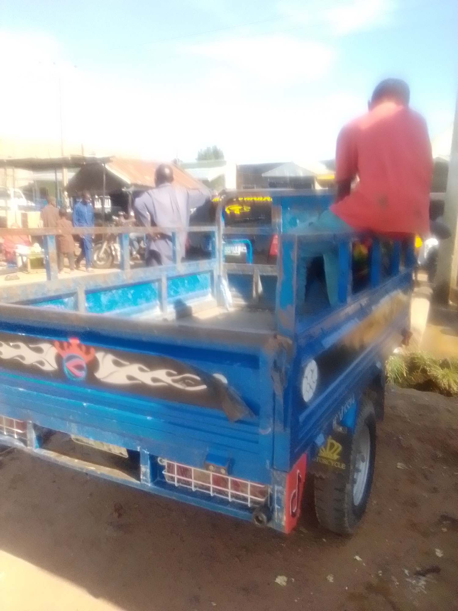 In Pantame, Nigeria, a blue three-wheeled vehicle is parked on a dirt road. The vehicle has a flatbed in the rear, which appears to be empty. A man wearing a red shirt is sitting on the edge of the flatbed, facing away from the camera. In the background, there are several market stalls with awnings and various goods displayed. A few people are visible near the stalls, some standing and some walking. There is also a motorcycle parked nearby. The sky is bright and appears to be clear, suggesting a sunny day. The overall scene depicts a typical market environment in Nigeria.