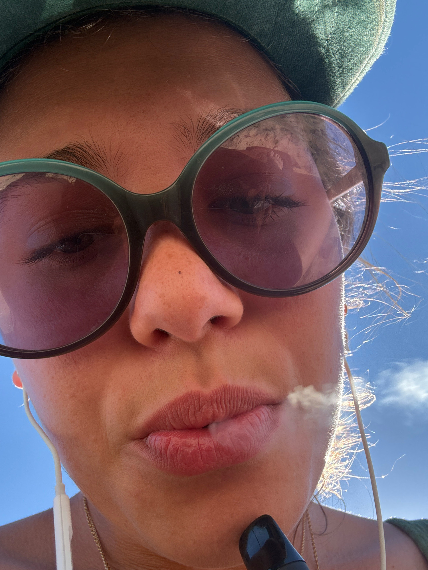 A close-up, low-angle shot captures a person wearing sunglasses and a green beret, against a clear blue sky. They are holding a dark object to their mouth, from which a thin stream of white vapor is emanating. The person has light brown skin, with visible pores and a few small dark spots. They are wearing white earbuds, with one visible wire extending towards the bottom of the frame. A delicate gold chain is also visible around their neck. The overall impression is a candid, outdoor moment, possibly during daytime given the bright sunlight and blue sky. The context of Sozopol, Bulgaria is not discernible from the visual information in the image.