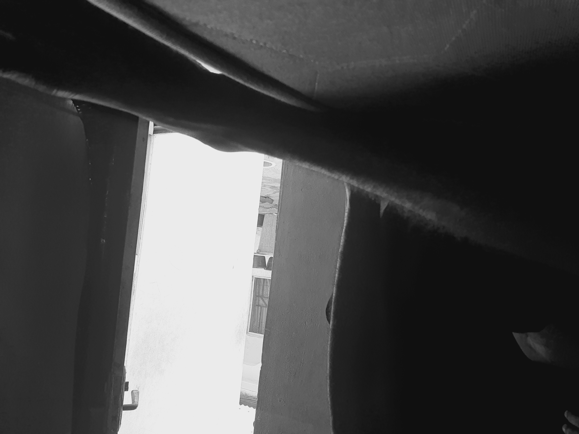 The image is a low-angle, black and white shot looking out from a dark interior space. The view is oriented toward a bright exterior scene, seemingly an urban landscape in Lokoja, Nigeria. In the background, there is a building with windows and what appears to be an air conditioning unit. The foreground is dominated by dark shapes, possibly parts of the room's ceiling or architectural details, obscuring the view partially. There are no visible people or actions in the frame. The environment suggests an enclosed space looking outward into a brightly lit area, possibly an open street or courtyard.