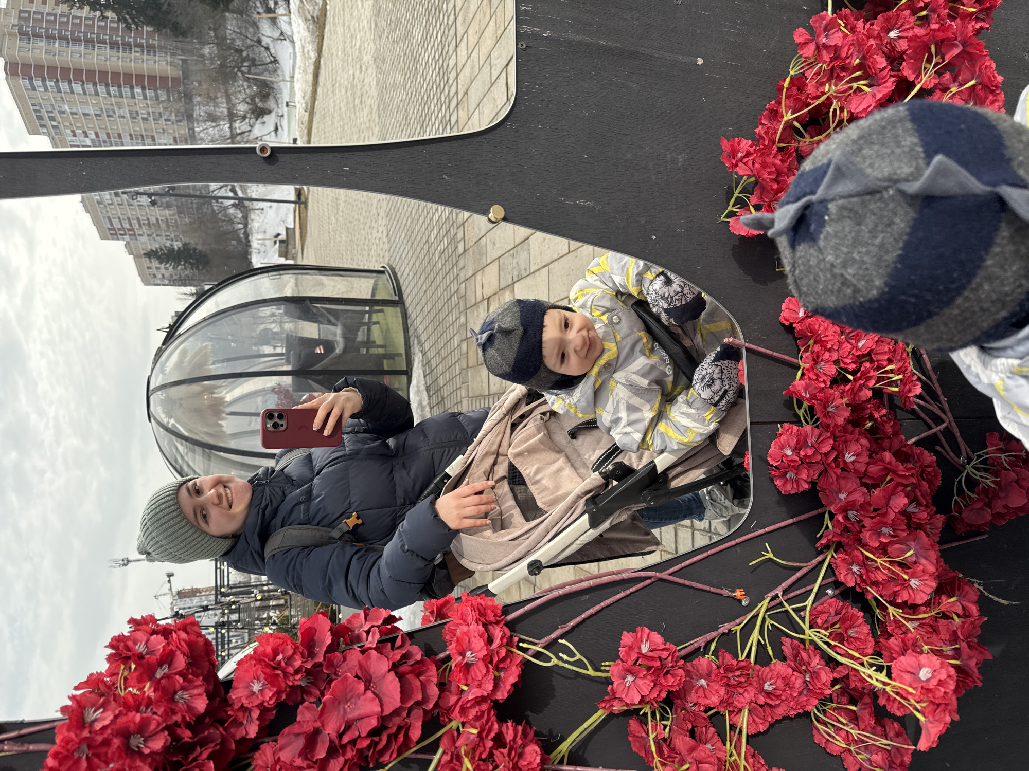 The image depicts an outdoor scene in an urban park or public space in Izhevsk, Russia. A woman, appearing to be an adult, is positioned on the left, smiling towards a red-cased smartphone held in her right hand, suggesting she is taking a photograph. She wears a grey knitted beanie, a dark blue puffer jacket with a backpack strap visible on her right shoulder, and blue jeans. Her left hand rests on the handle of a light beige baby stroller.
Seated in the stroller is a child, identified as a toddler or infant, wearing a grey and dark blue striped knitted hat with a dorsal fin-like element, a grey and yellow patterned winter jumpsuit, and patterned mittens. The child is looking slightly to the left of the camera with a faint smile.
The right side and lower left foreground are occupied by a dark, irregularly shaped panel or decorative mirror frame. This surface is extensively adorned with clusters of artificial red flowers featuring green and yellow stems. The woman and child are reflected within this panel, with the child's reflection occupying the lower central part of the mirrored area.
In the mid-ground, behind the subjects, are several clear, geodesic dome-shaped outdoor structures, one of which contains seating. The ground in this area is paved with light-colored interlocking blocks. The background features a sloped terrain covered in patches of snow, with bare deciduous trees and evergreen trees. Multi-story residential buildings with beige, red, and brown facades are visible on higher ground behind the trees. A construction crane is faintly discernible in the far background to the left. The sky is overcast, indicating daytime conditions during a cold season.