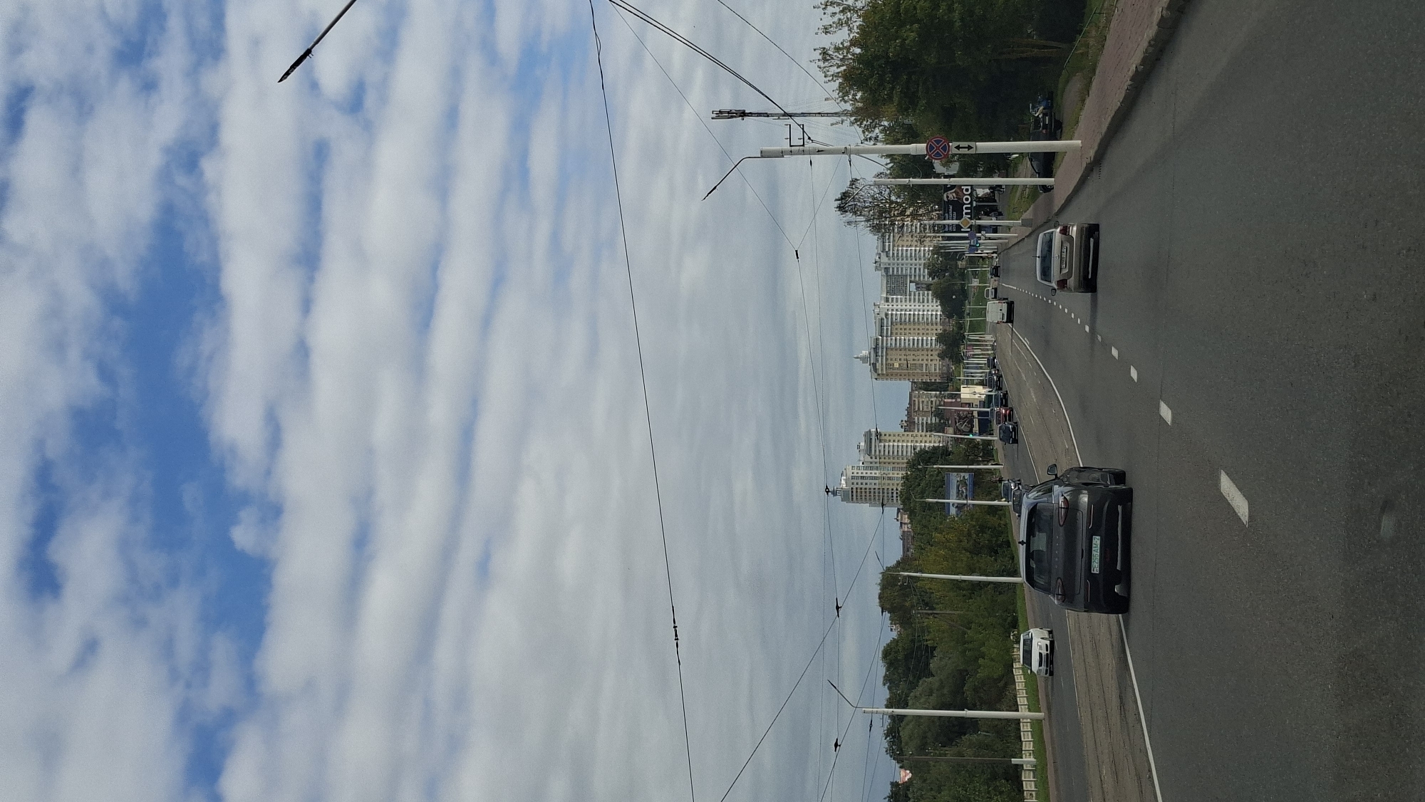The image depicts a view of a multi-lane highway in Vitsebsk, Belarus, with modern high-rise buildings in the distance. The sky is partly cloudy with a mix of blue sky and white clouds. The highway has clear white lane markings and is populated with several vehicles, including cars and a van. Trees line the sides of the road, and there are streetlights and utility poles visible. The perspective is from a vehicle traveling on the highway.