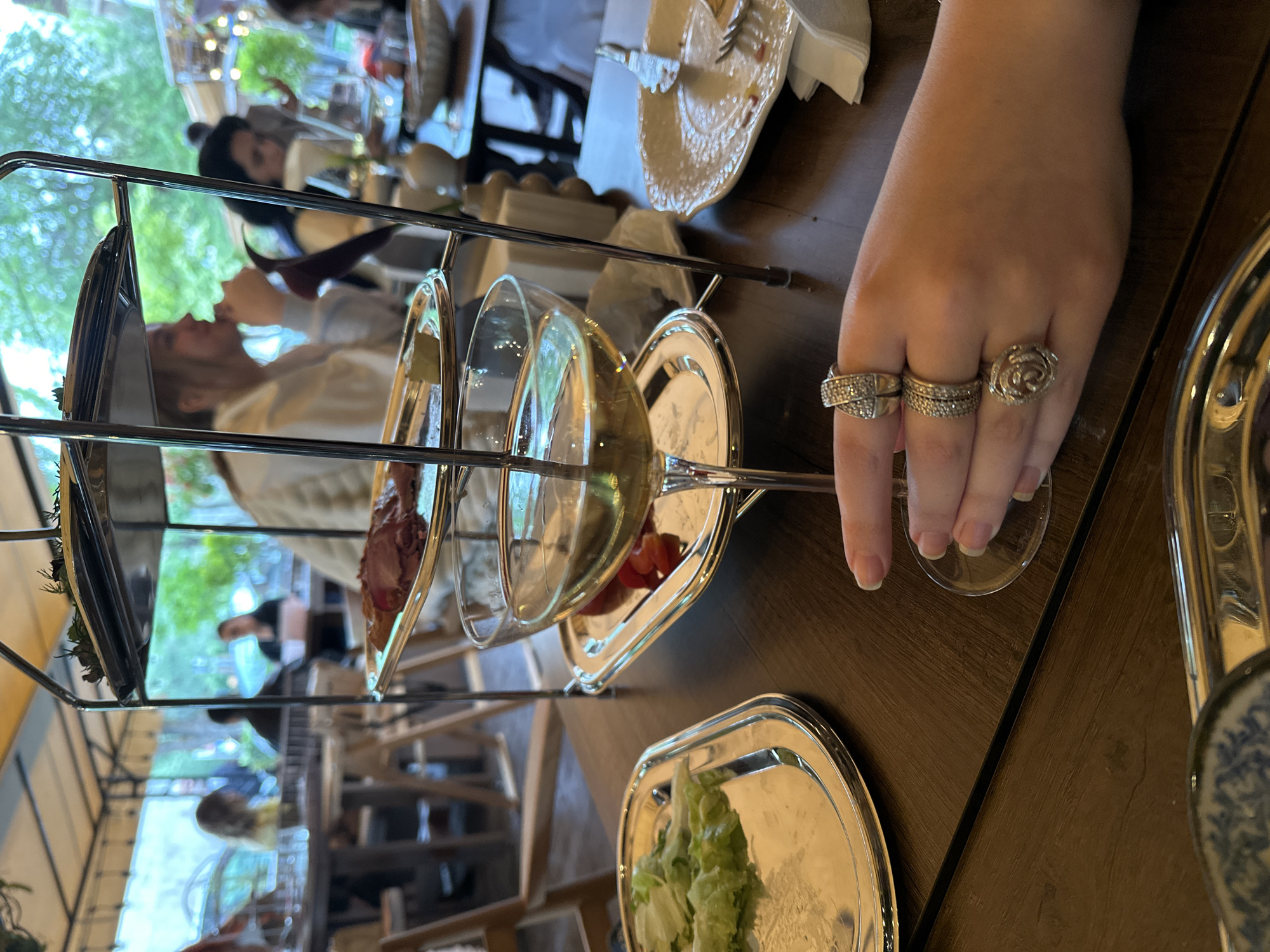 The image depicts a dining scene in Yerevan, Armenia. A woman's hand, adorned with three rings, holds a wine glass. A three-tiered serving tray holds food items. Other people are present at nearby tables.