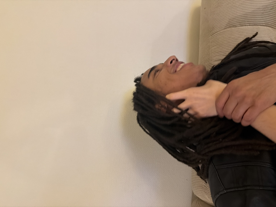 An indoor scene depicts two individuals in a close interaction. One individual, appearing to be a male with dark skin and long dark dreadlocks, is seated on a beige, ribbed sofa, facing right in profile, smiling. He wears a black leather-style jacket. His right hand grasps the left forearm of a second individual, who has lighter skin. The second individual's left hand is placed gently on the first individual's neck/jawline area. The background consists of a plain, light-colored wall. The exact location within Paris, France, cannot be determined from the visual information provided.