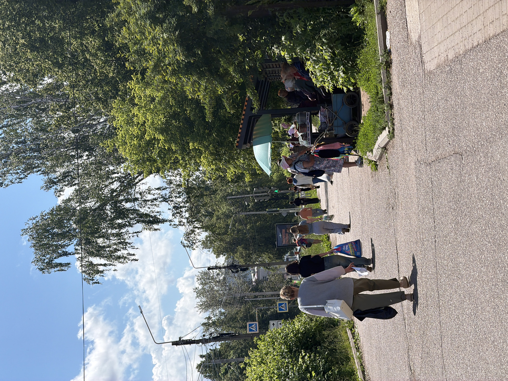 A sunny day in Chaikovsky, Russia, depicts a street scene with people walking and a small outdoor market. In the foreground, two individuals are walking away from the camera on a paved street. The person on the left is wearing a grey cardigan and green pants, carrying a white purse and a black bag. The person to their right wears a black top and blue jeans, holding a blue shopping bag. Further down the street, more people are walking, some with shopping bags.
To the right of the street, a covered stall with a green umbrella is set up, where several people are gathered, appearing to be customers or vendors. Items are displayed on a cart in front of the stall. Lush green trees and bushes surround the area, with streetlights and power lines visible against a blue sky with scattered white clouds. The ground is asphalt with a sidewalk or paved path to the right. The overall atmosphere suggests a casual day in a suburban or town setting.