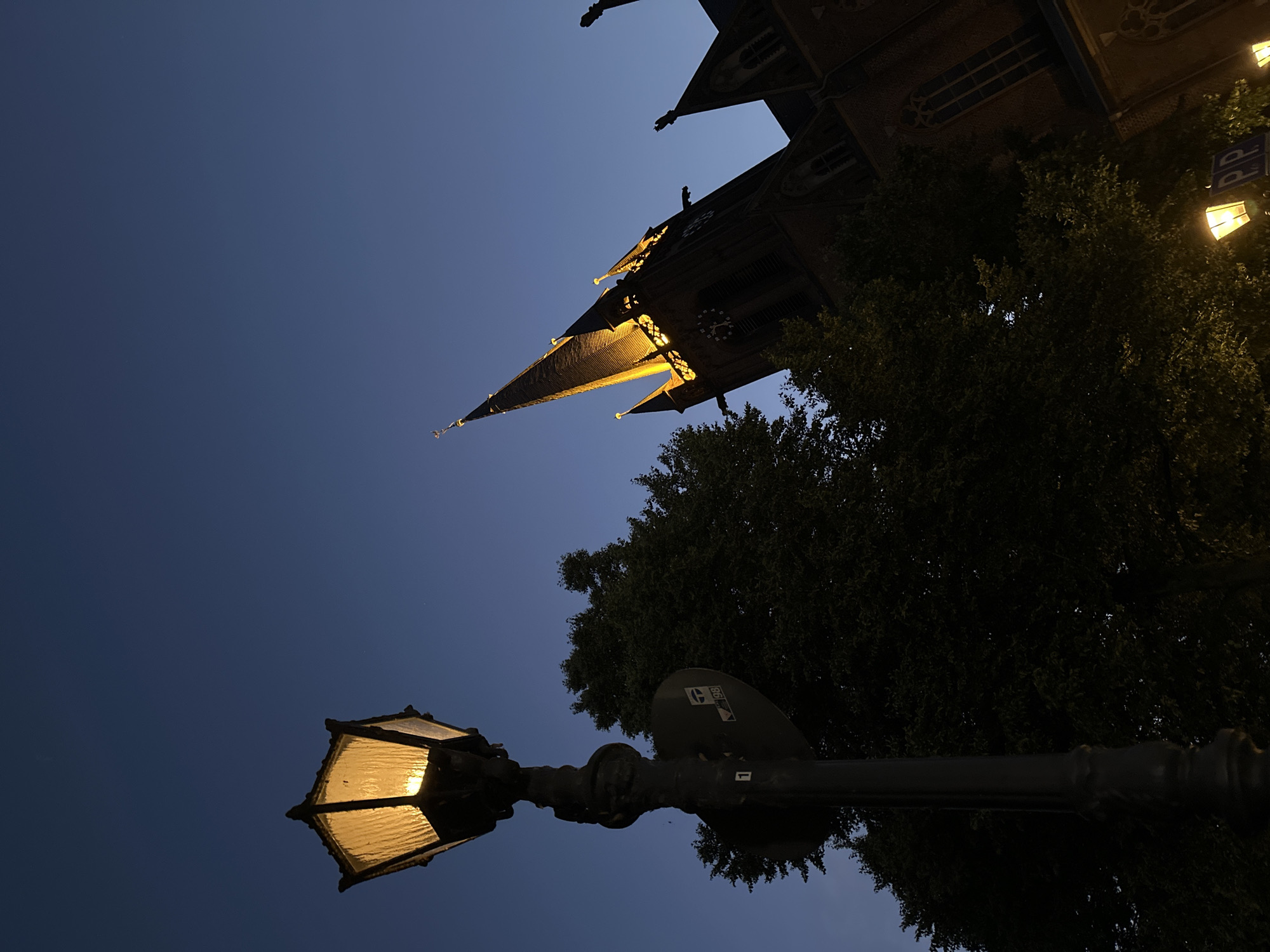 The image depicts a low-angle view of a street lamp and a church spire against a deep blue twilight sky. The street lamp, in the foreground on the left, is illuminated with a warm yellow light. It is mounted on a dark, ornate pole which has a round sign attached to it, displaying white text and symbols, including the number '1' and a graphic with '98' visible. Behind the lamp post and to the right, the dark silhouette of a large, leafy tree obscures much of the lower portion of the scene. Emerging from behind the tree and dominating the right side of the image is the spire and upper facade of a church. The church is constructed of dark brick, and its spire and decorative elements are brightly illuminated with a warm yellow light, highlighting its gothic architecture. Several gargoyles are visible on the church's exterior. The sky is clear and a gradient from deep blue at the top to a lighter shade near the horizon. Two additional illuminated street lamps are visible in the bottom right corner of the image. This scene is likely located in Krefeld, Germany, given the specific signage and architectural style, potentially near the Stadtwald.