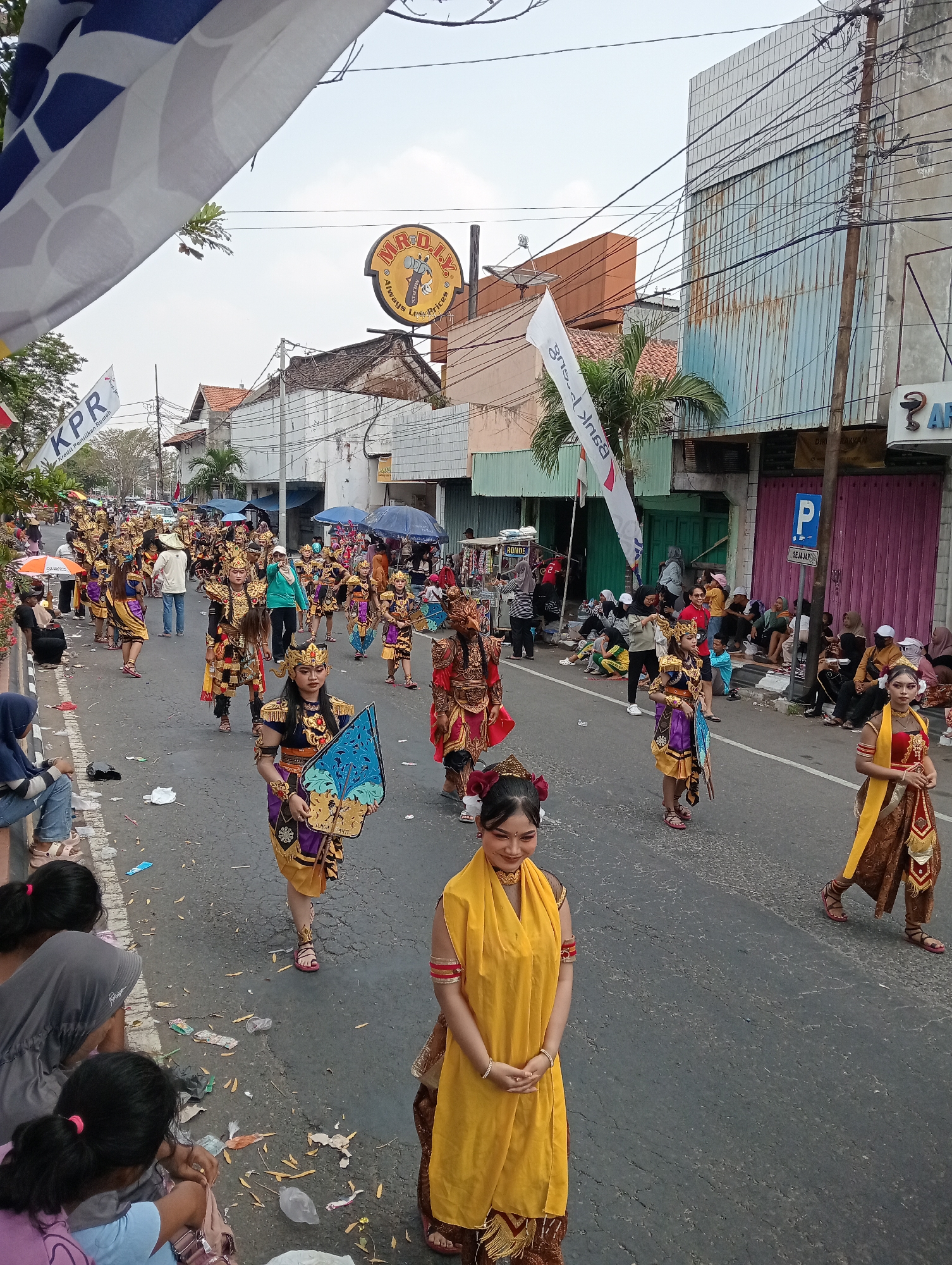 This is a full, high-angle shot of a parade in the city of Demak, Indonesia. A group of people in traditional Javanese costumes, elaborate headdresses, and face paint are walking down a paved street lined with buildings and spectators. The performers are adorned with intricate gold and colorful fabrics, some carrying decorative fans or shields. The street is littered with trash, including plastic bottles and wrappers, indicating a festive or public event. Spectators are seated on the roadside and standing, some holding umbrellas for shade. Flags, including one with "KPR" printed on it and another with a "MR. D.I.Y." logo, are visible. The buildings appear to be a mix of commercial establishments and residential structures, with visible electrical wires and utility poles. The overall scene depicts a vibrant cultural celebration in a bustling urban environment.