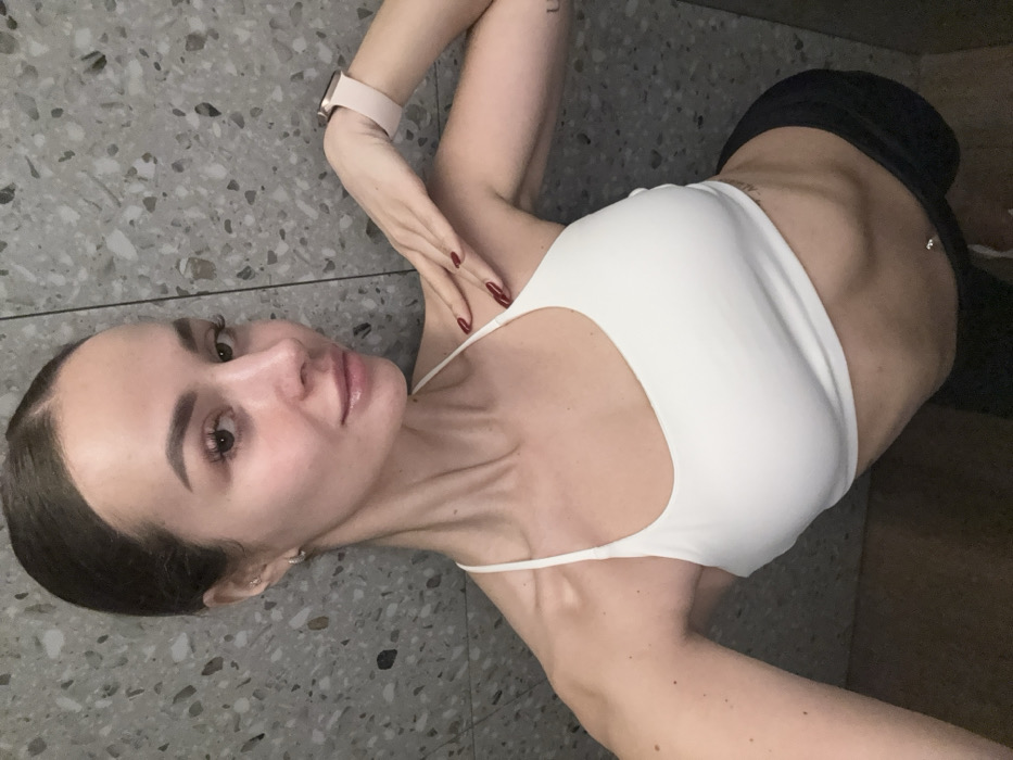 A single adult female is depicted in an indoor selfie, with the scene located in the city of Kyiv, Ukraine. She has dark hair pulled back and wears a white athletic bra and black lower garments. Visible accessories include small silver-colored hoop earrings, a light pink smartwatch on her left wrist, and red nail polish on her fingernails. A silver-colored barbell navel piercing is present, along with a partial dark-letter tattoo on her left upper arm. The subject's left hand is positioned on her right shoulder/neck area. The background consists of a uniform grey wall surface with a speckled, terrazzo-like pattern.