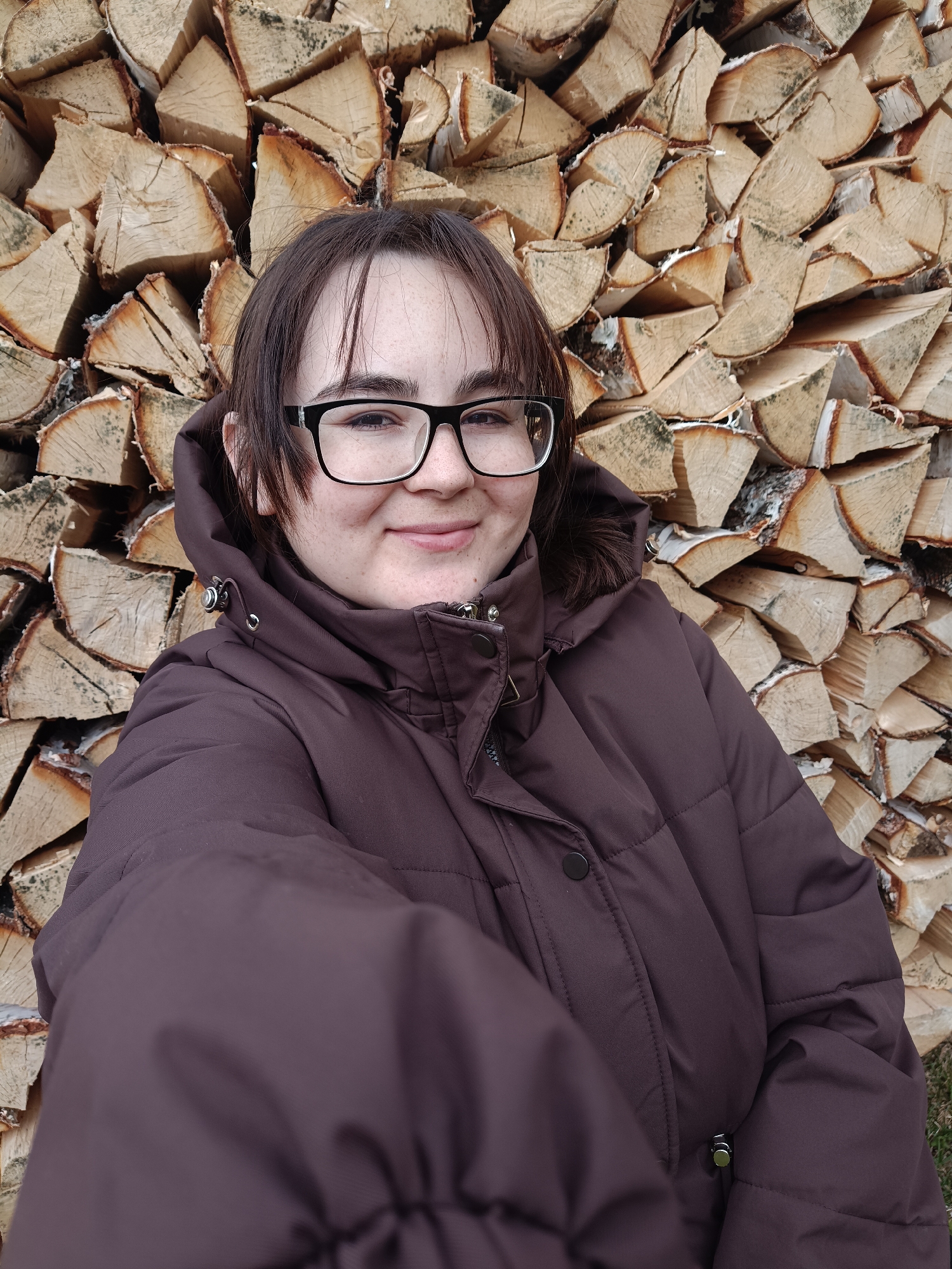 A young person, likely female, with dark hair and black-framed glasses, is featured in a close-up, selfie-style photograph. She wears a dark brown, quilted winter coat with a hood. Her expression is a gentle smile, conveying contentment. Behind her, a substantial stack of neatly piled firewood dominates the background. The logs appear to be birch, characterized by their light, often peeling bark, and show clear signs of being split and seasoned. This setting, with the person dressed for cold and the prominent firewood stack, is consistent with the provided location context of Barda, Russia, where such wood is commonly used for heating. The natural lighting suggests it is daytime. There is no visible text in the image.