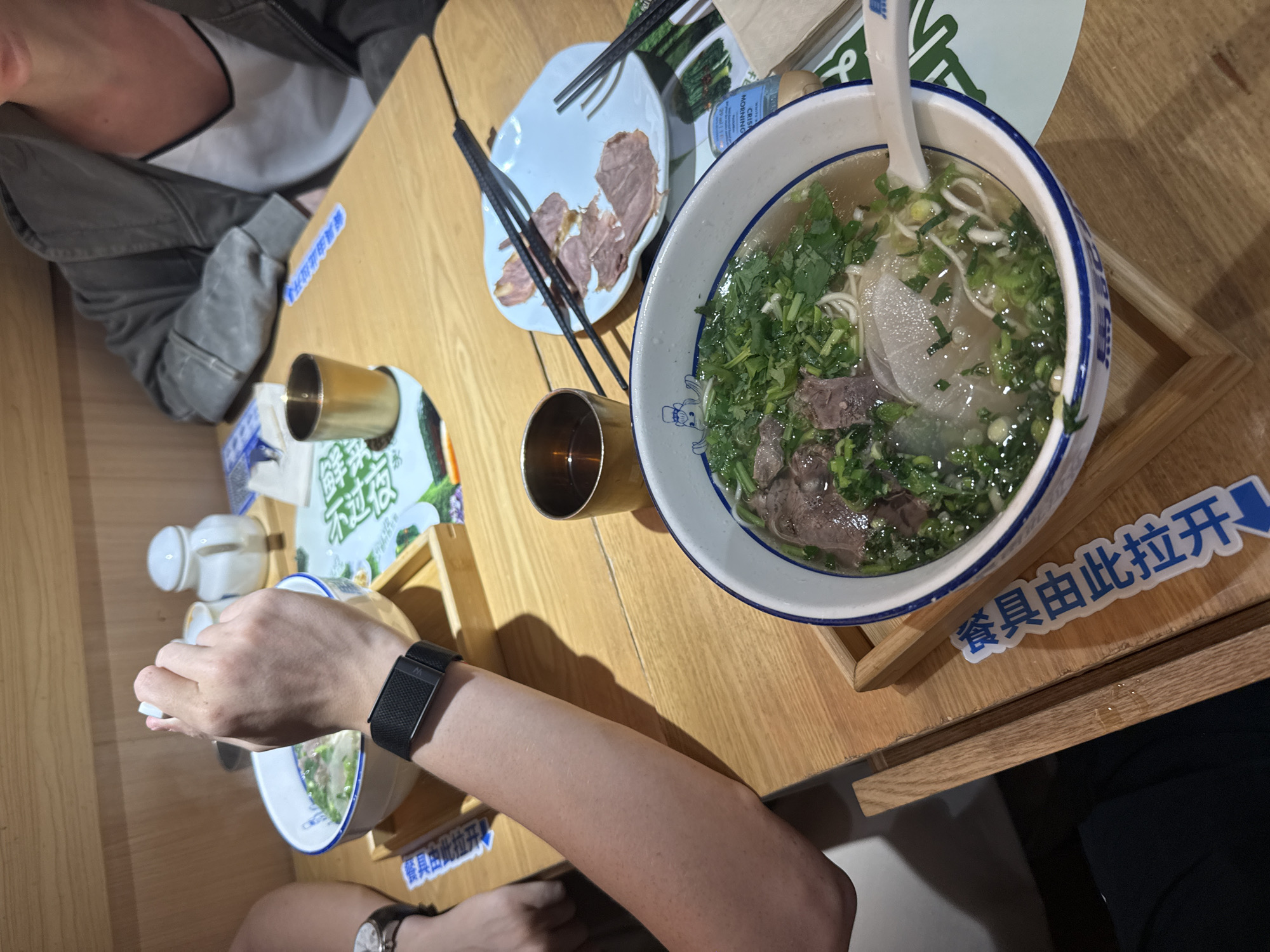The image shows a person at a table in a restaurant setting in Tianhe District, China. The table holds a bowl of soup with noodles, meat, and green garnish. A small plate with pieces of meat and chopsticks is also visible. A small metallic cup is also present, along with what appear to be condiments. The person is wearing a black smart watch on their left wrist and a watch on the other. The environment suggests an indoor dining area with light wooden surfaces.