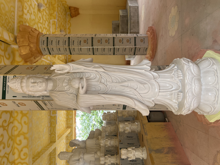 A large white marble statue of a standing female deity, likely Quan Âm (Guanyin), occupies the foreground of an indoor or semi-enclosed space in Đăk Mar, Vietnam. The statue features a crown, flowing robes, a beaded necklace, and holds a lotus bud in its left hand while its right hand is raised in a mudra. It rests on a multi-tiered lotus pedestal.
To the left of the main statue, a collection of smaller white marble or stone statues are arranged on shelves and directly on the floor. These include seated figures, some appearing to be wrapped in protective plastic, and several pairs of standing figures.
The environment exhibits characteristics of a construction or renovation site. Several structural pillars are prominently visible, wrapped in protective white panels branded with "Gia Phát" and "ALUMINUM COMPOSITE PANEL." The ceiling is painted a bright yellow with intricate, swirling patterns, and a large golden lotus-shaped capital adorns one pillar. The floor is a light-colored, possibly concrete, surface with faded circular designs. In the background right, a set of unadorned concrete steps leads upwards. Green foliage is visible through an opening on the left, indicating an external view.