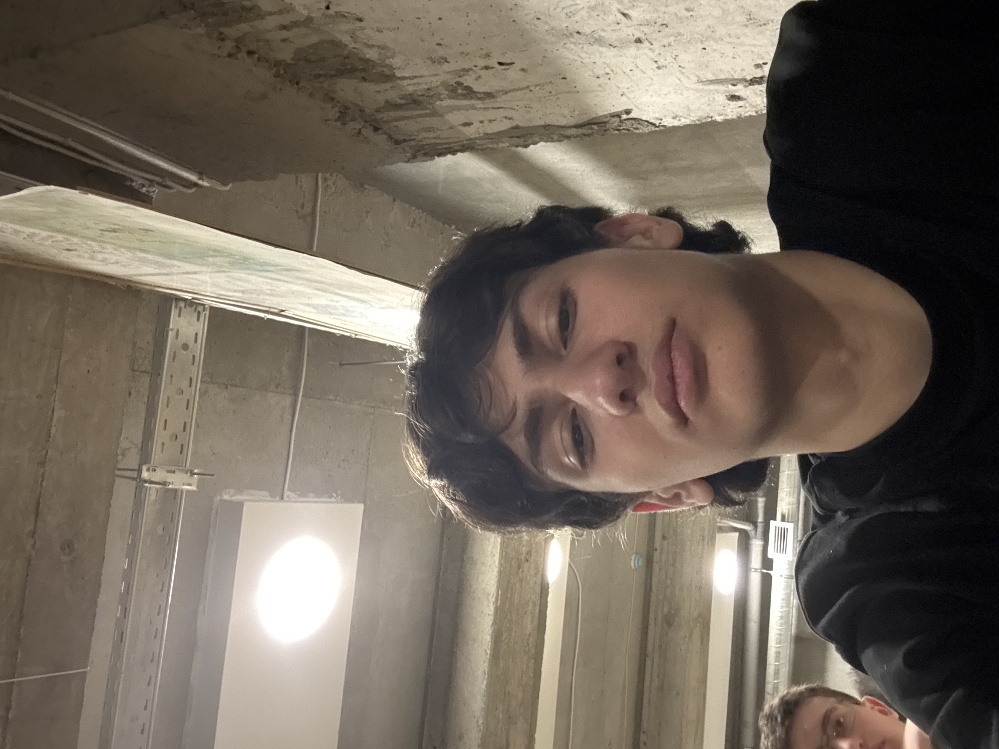 The image appears to be taken in an unfinished or industrial-looking concrete room or garage. A young man with dark hair and a black shirt is in the foreground, holding the camera at an upward angle, capturing his face and the ceiling. The ceiling is bare concrete with exposed pipes and electrical conduit. There are fluorescent lights and metal structural supports along the walls. In the bottom right corner, a partial view of another person's face is visible. There is no information available to establish the location in Yerevan, Armenia.