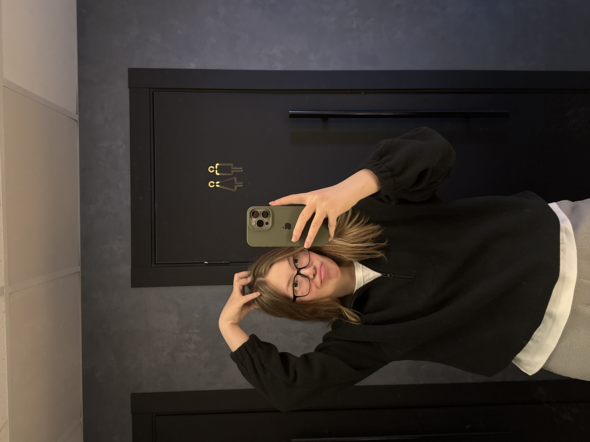 A young woman is taking a selfie in front of a dark-colored door in a restroom. The door has gold-colored restroom signage. The woman is wearing glasses and a dark-colored, long-sleeved, zip-up fleece sweater over a white shirt. She is holding an olive-green iPhone. The background includes a light-colored wall. There is no indication of the city of Chaikovsky, Russia, in the image.