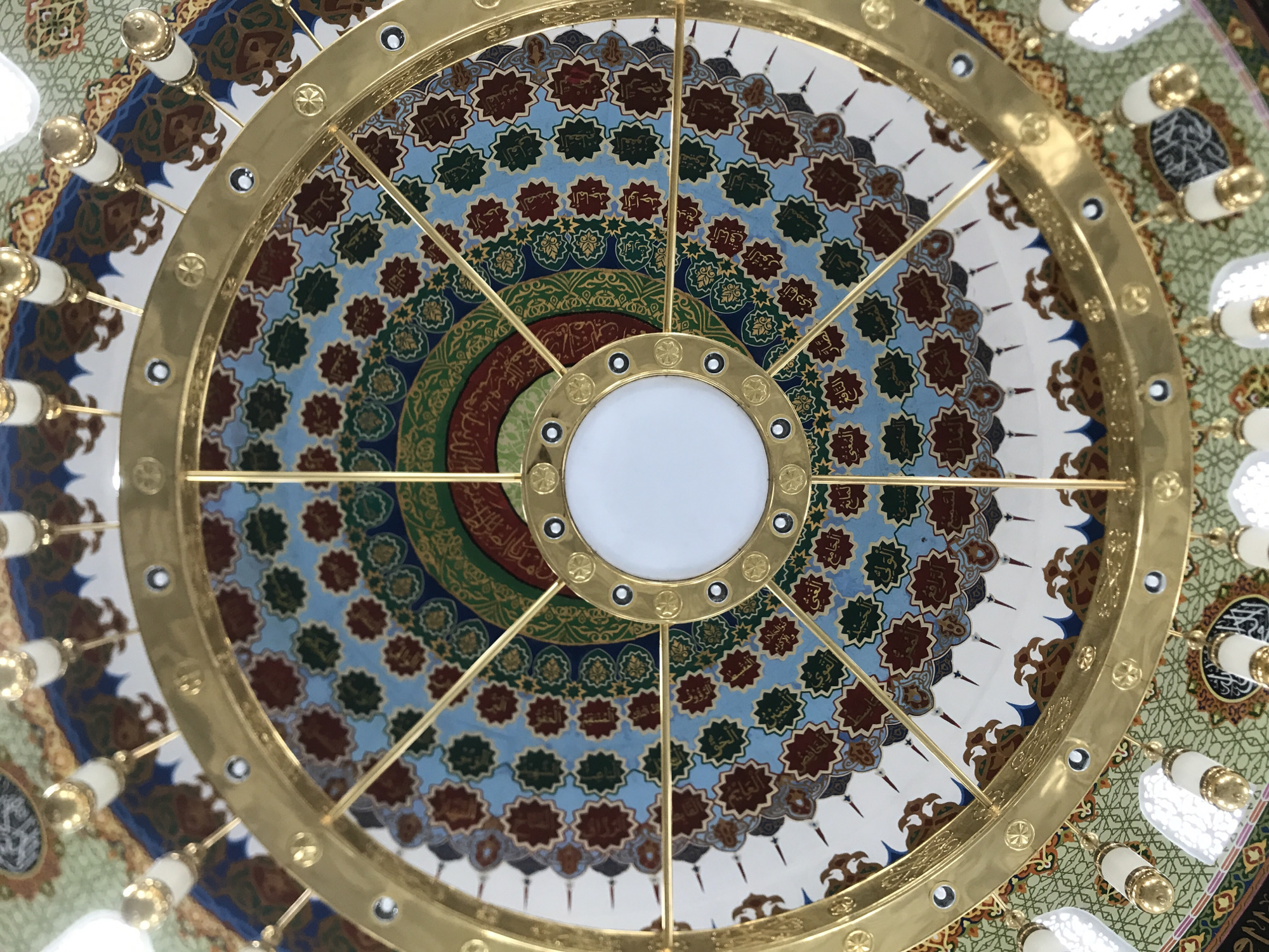 A low-angle, eye-level shot captures a circular, golden chandelier set against a decorative ceiling. The chandelier features a central white light surrounded by a series of concentric golden rings connected by radial rods. Decorative light fixtures are attached to the outer ring. The ceiling behind the chandelier is patterned with geometric designs and Arabic calligraphy in blue, green, brown, and gold. The image was likely taken inside a mosque. The location is Meunasahaluedrienlhoksukon Barat, Indonesia.