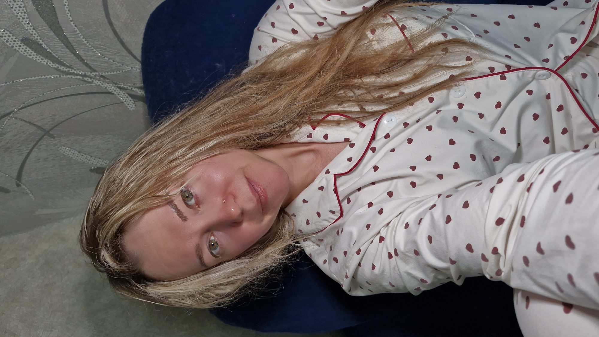 A woman is lying on a dark blue cushion, angled to the left. She is wearing cream-colored pajamas with small red hearts and red piping along the collar and front opening. The pajamas are buttoned at the top. The woman has fair skin, light blonde hair, and green eyes. The background shows a white surface with a patterned design, and part of a light-colored rug. The specific location in the city of Otchor, Russia cannot be determined from the image.