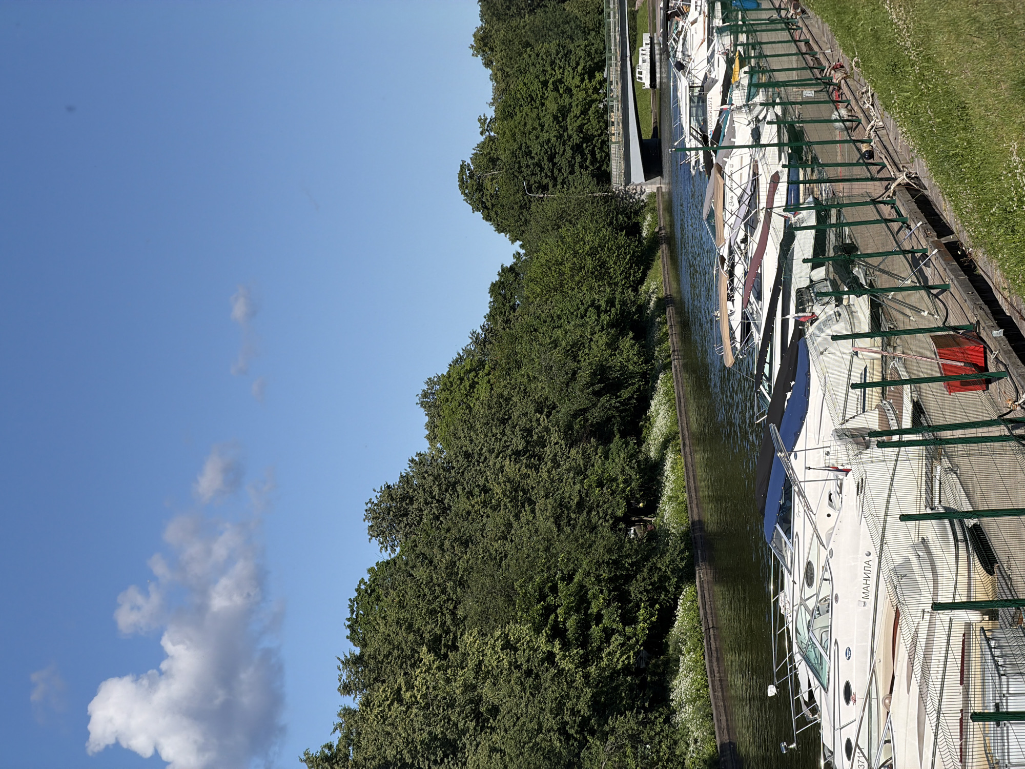 The image displays a canal with several yachts docked along the side. The canal is lined with lush green trees and grass. The sky is a clear blue with a few scattered clouds, suggesting a pleasant day. The yachts are white with blue and tan accents, and some have awnings. One yacht has the name "МАНИЛА" visible on its side. The scene appears to be in a marina or a canal within a city, with a fence separating the water from the grassy area. The orientation of the image is vertical, and the scene is viewed from a slightly elevated perspective. The time of day appears to be daytime, likely mid-morning or afternoon, given the bright sunlight and clear sky. The overall atmosphere is peaceful and serene.