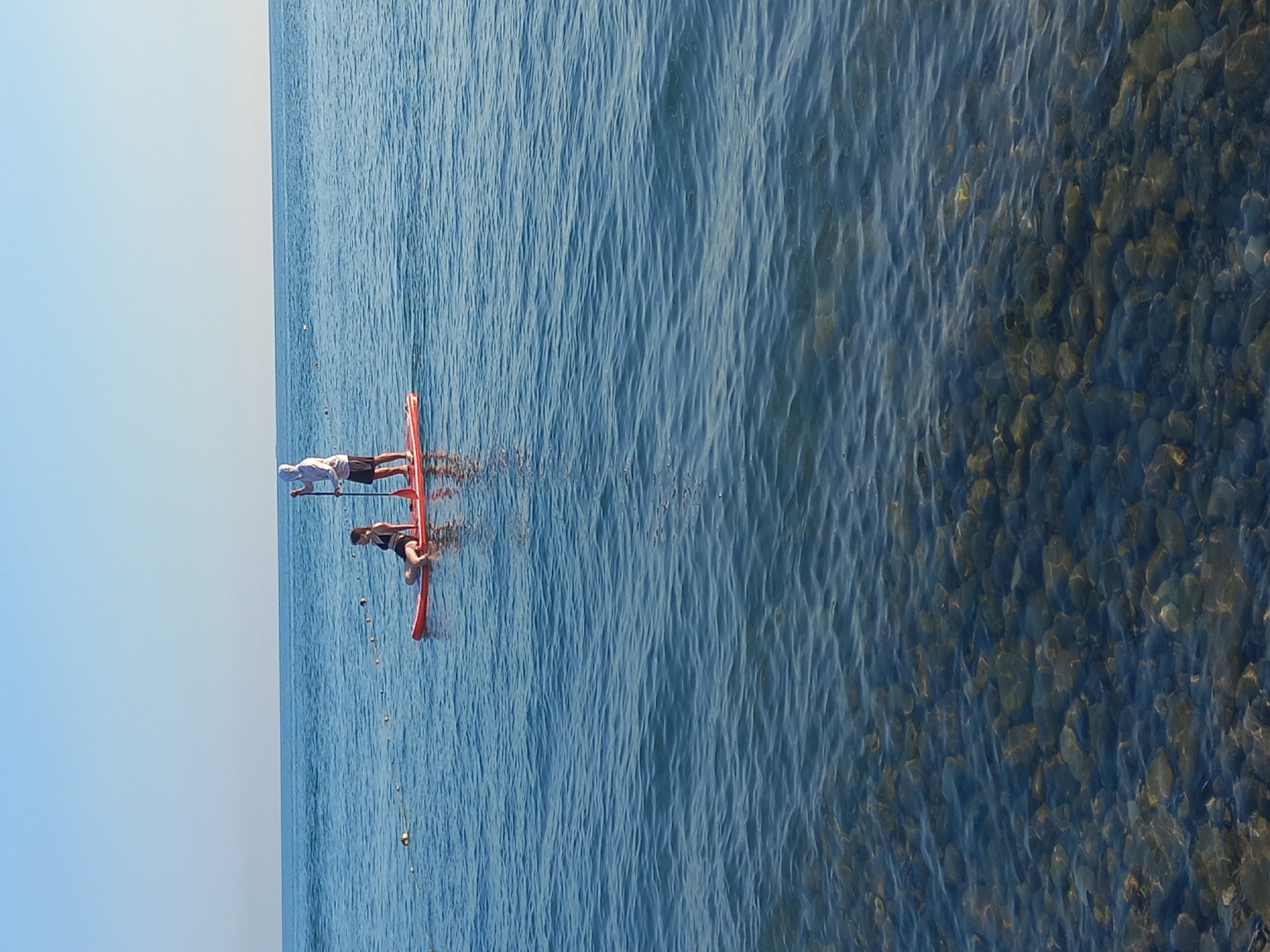 Two people are paddleboarding on the Black Sea near Sochi, Russia. The person in the front is a woman wearing a black bikini, sitting on the orange paddleboard. The person behind her is a man wearing a white hooded jacket and dark shorts, standing and holding a paddle. The water is calm with ripples, and the seabed with pebbles is visible through the clear water in the foreground. The sky is clear and blue.