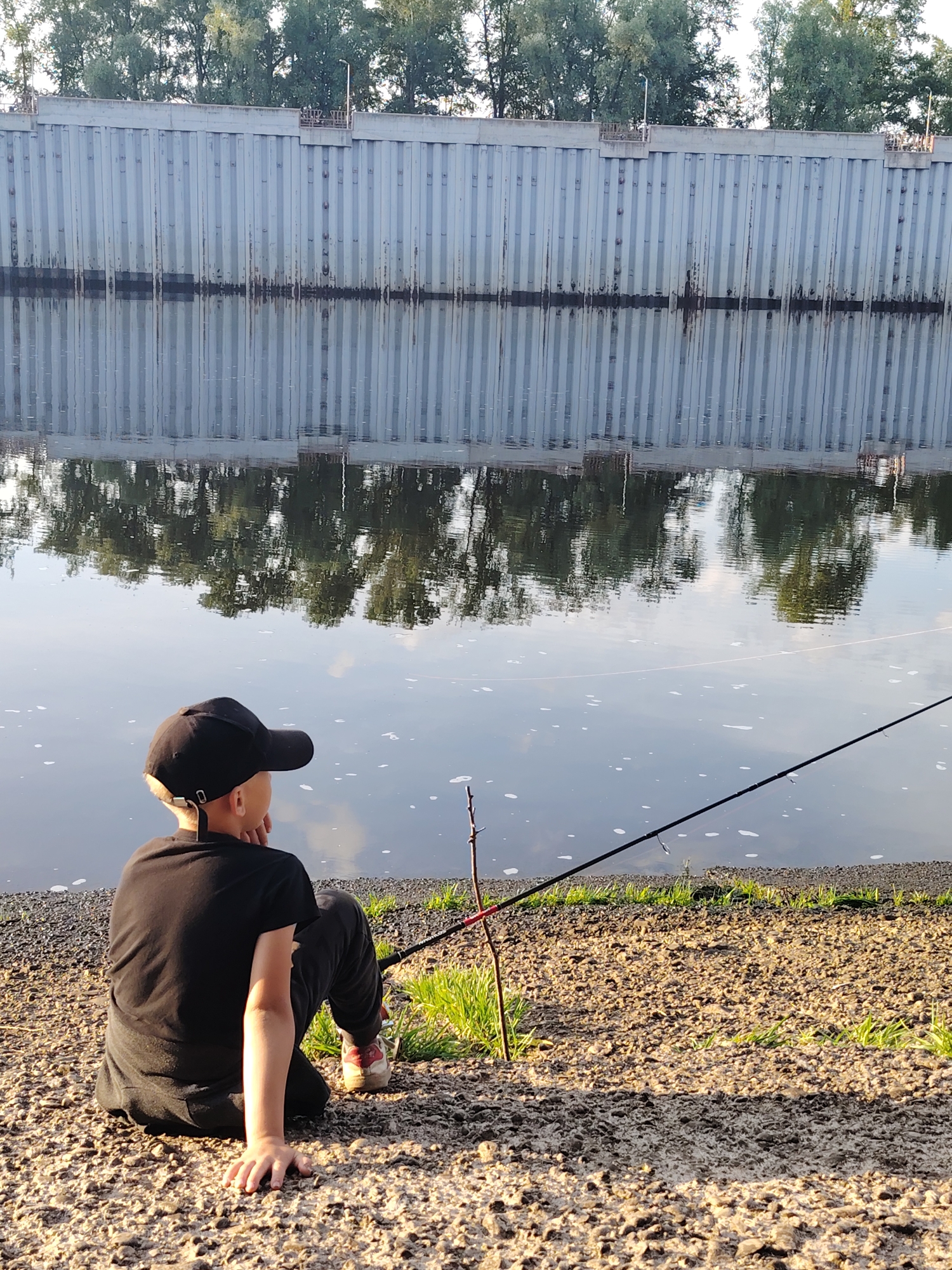 A young boy wearing a black t-shirt and pants, and a black baseball cap, sits on a gravelly shore facing a river, with a fishing rod in front of him. The river has a reflective surface showing the reflection of a tall, vertical metal wall on the opposite bank, and trees above it. The wall appears to be a flood defense or embankment. The scene is located along the Kama River in Chaikovsky, Russia. The sun is casting long shadows, indicating it is likely late afternoon or early evening.