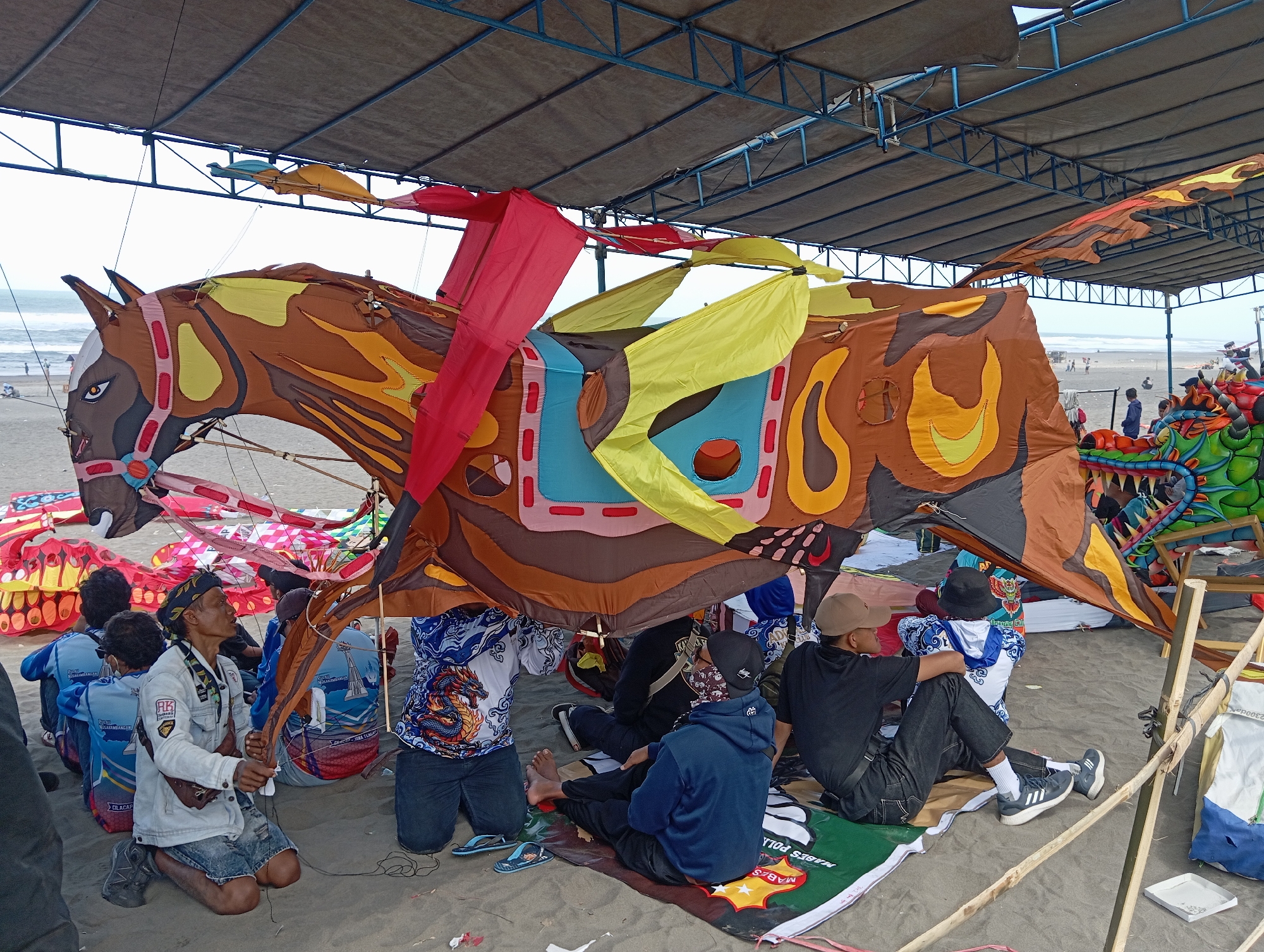 In Parangtritis, Indonesia, a group of people are gathered on a sandy beach under a large, shaded canopy. The central focus is a massive, colorful kite in the shape of a brown and orange horse with red and yellow accents, its head facing left. Several other large kites, including a green dragon, are also visible. The people, some kneeling and some sitting, are interacting with the kites. One person in the foreground, a man wearing a light-colored jacket and denim shorts, is kneeling and holding kite strings. Others are seated around the large horse kite, some looking towards it. The environment is a beach with the ocean visible in the background, and the sky is overcast. The overall scene suggests a kite festival or a preparation for one.