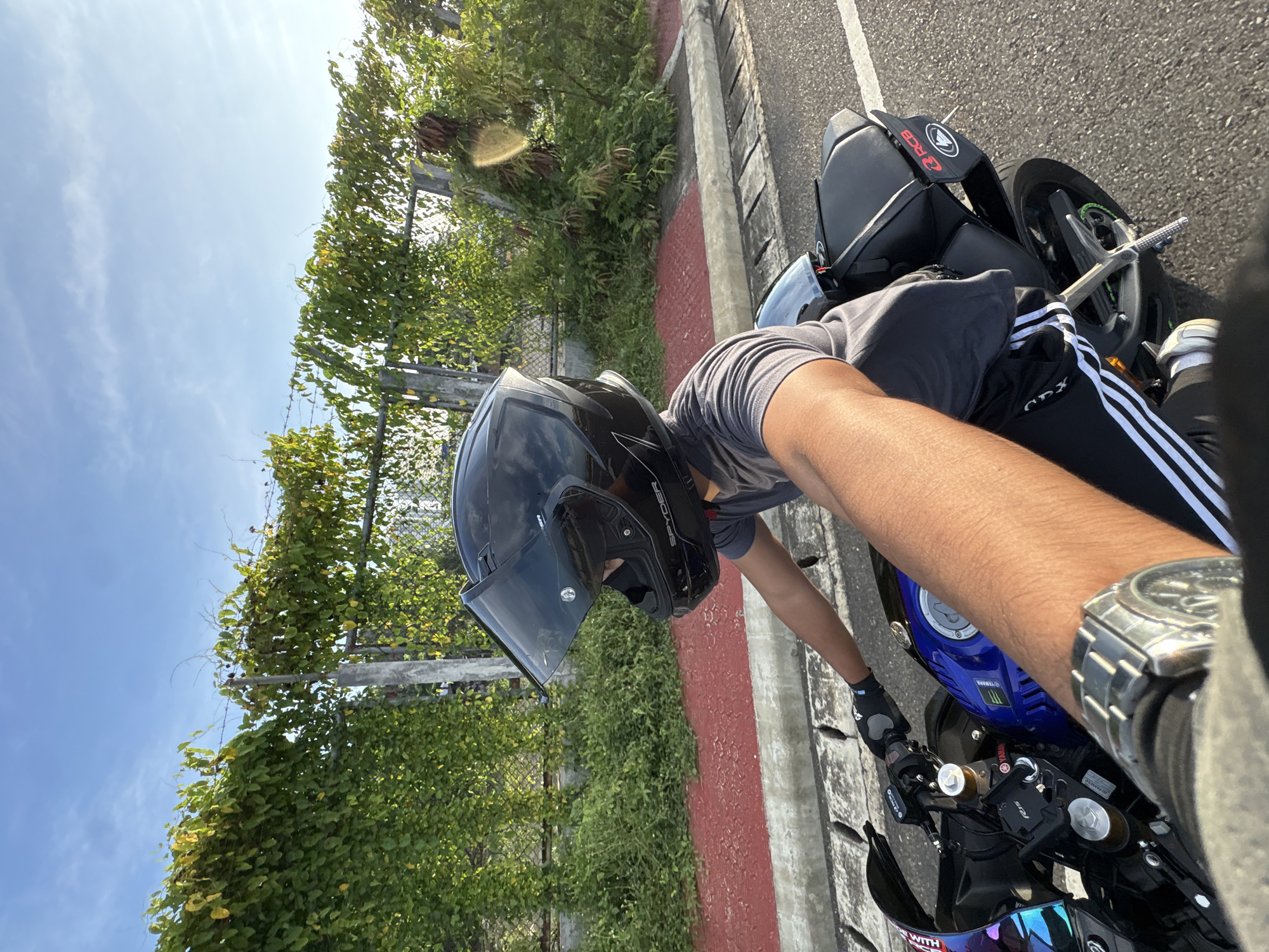 The image depicts a person riding a blue motorcycle, viewed from a first-person perspective. The rider is wearing a black full-face helmet and a grey t-shirt. They are also wearing black gloves and black athletic pants with white stripes. A silver watch is visible on the rider's left wrist. The motorcycle is a sportbike with the brand "Yamaha" visible on its fuel tank. A black bag is attached to the rear of the motorcycle. The background shows a roadside with a concrete curb and a red-colored paved area. Beyond the curb, there is a dense green vine-covered fence or wall, with a wire fence visible through the foliage. Green bushes and small trees are also present. The sky is clear blue with a few wispy clouds. The lighting suggests it is daytime, likely late afternoon due to the warm tones of the sunlight. Based on the context of the photographer’s location (Davao City, Philippines), this scene likely occurs along a road in or near the city, possibly a less developed or more scenic area with natural vegetation bordering the road.