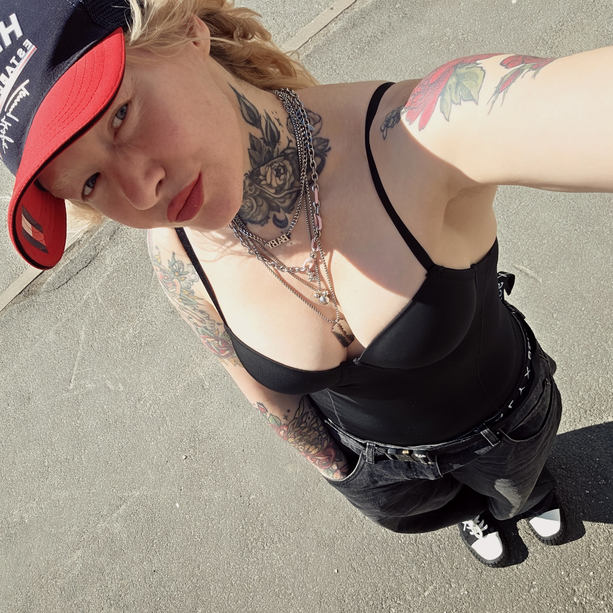 A fair-skinned person wearing a red and blue baseball cap, black top, black jeans with a white logo band, and black and white shoes is standing on a gray concrete surface, possibly pavement, in direct sunlight. The person has multiple tattoos on their arms and neck, and wears multiple silver-colored necklaces with different pendants. The person's posture suggests they are taking a selfie from a high angle. The setting appears to be an outdoor urban area. It is not possible to confirm the location in Saint Petersburg, Russia based on the provided image alone.