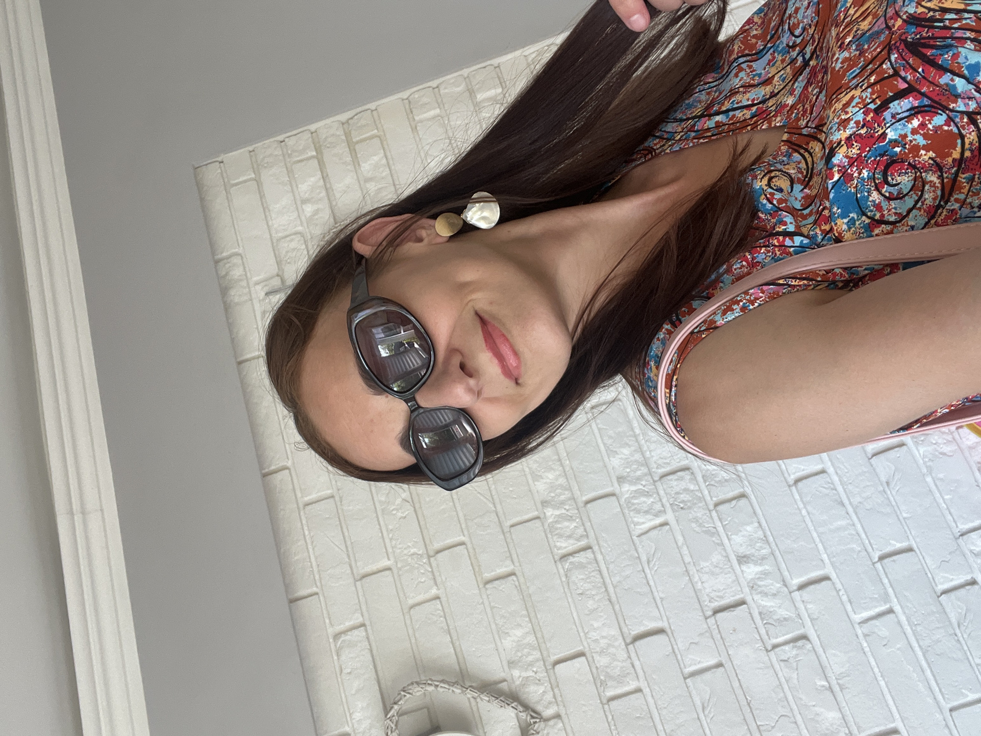 A woman with long brown hair is photographed from a low angle, her head tilted upwards and to the right. She is wearing sunglasses with dark lenses and a patterned dress with vibrant colors including red, blue, and yellow. Two earrings are visible on her left ear; one is a small gold stud, and the other is a larger, irregular-shaped pearl or iridescent earring. Her lips are painted pink, and she appears to be smiling slightly. A portion of a light pink purse strap is visible over her right shoulder. The background consists of a white brick-patterned wall and a white decorative molding on the left side of the frame, suggesting an indoor setting. The lighting is bright, indicating it could be daytime, but the specific time of day or weather conditions are not discernible. There is no visible text in the image.