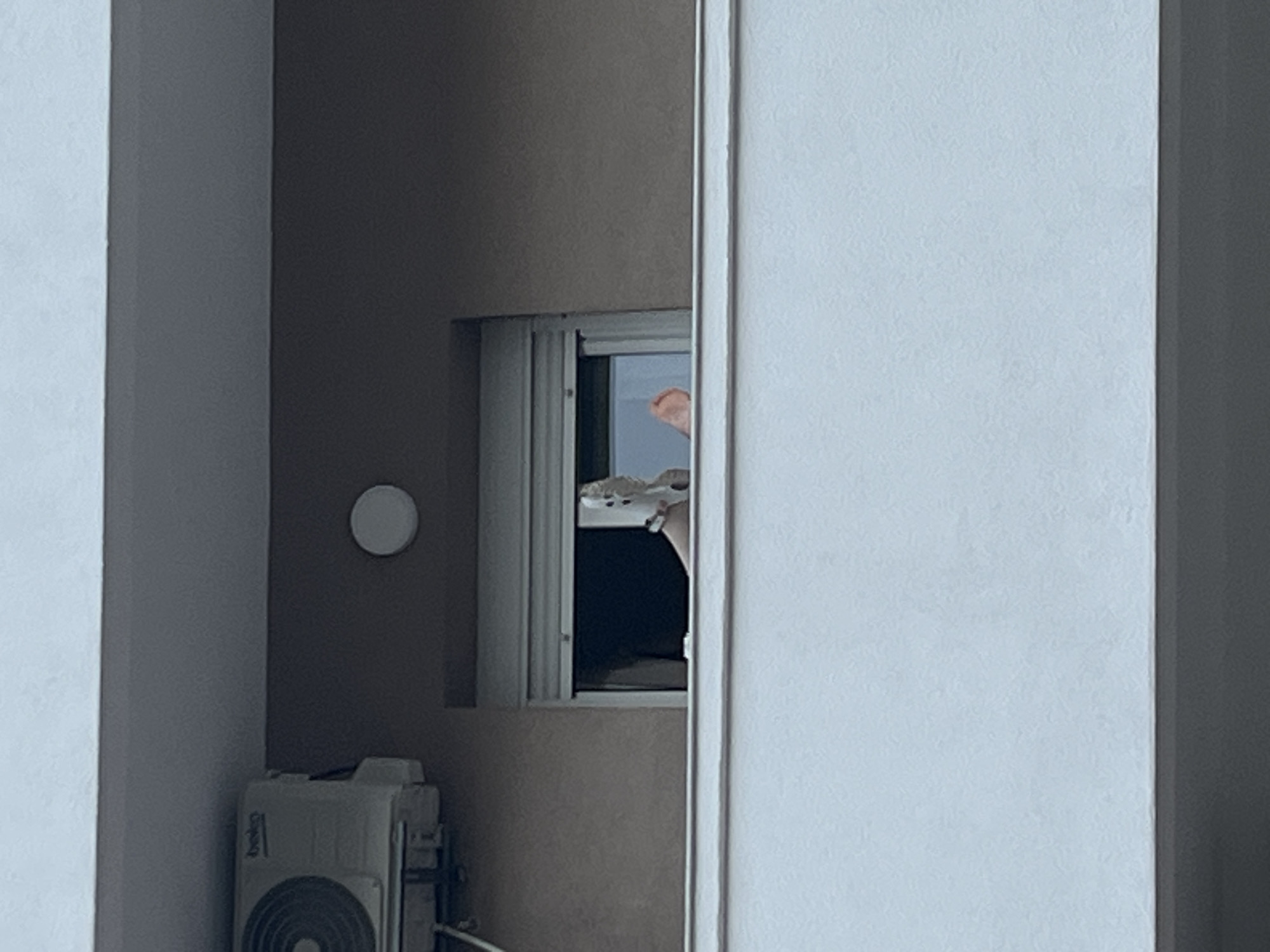In Budva, Montenegro, a person's legs are visible protruding from a window of a building. The left leg is higher and appears to be wearing a white sandal. The right leg is lower and the foot is bare, with the toes visible. The window has a white roller blind pulled down to about halfway. An air conditioning unit is mounted on the exterior wall of the building to the left of the window. The building has a light-colored balcony railing in the foreground. The sky is a light, overcast gray.