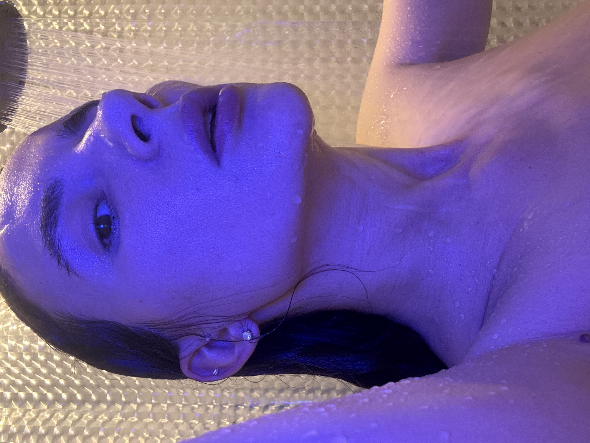 A woman lies in a shower illuminated by blue light. Water streams from the shower head onto her face and body. She is lying on a patterned shower floor. She has dark hair and an earring. The setting is a bathroom. It's not possible to discern if this is in Chaikovsky, Russia or any other location.