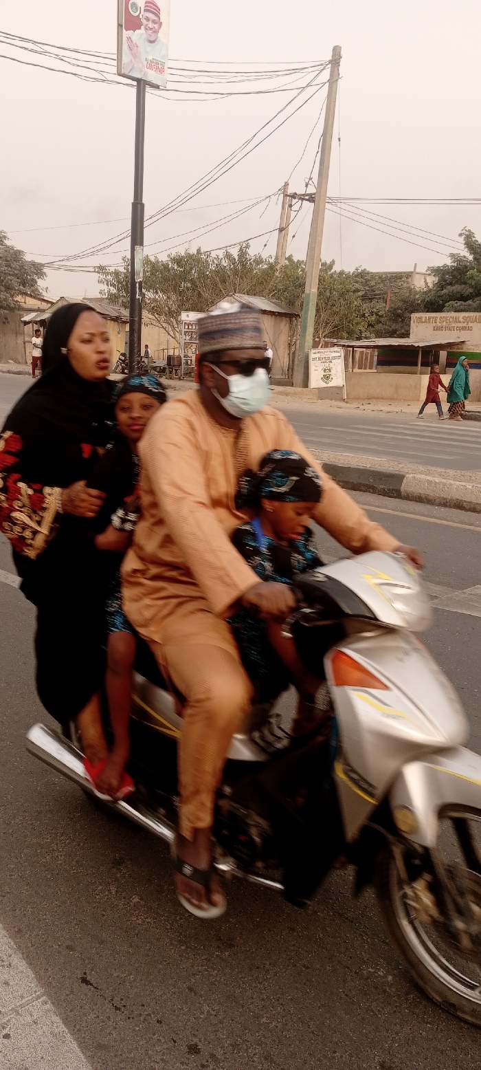 A man wearing a face mask and hat rides a silver motorcycle on a street. He is followed by two children and a woman. The woman wears a black hijab with floral patterns. A sign is visible on a pole in the background. Buildings and people walking can be seen in the distance on either side of the road.