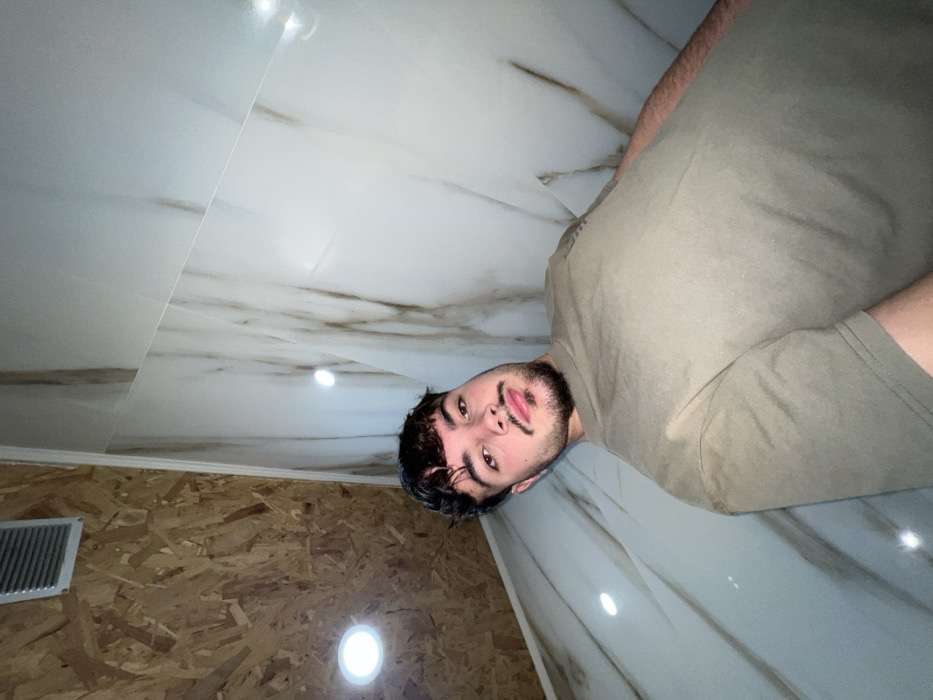 The scene, located in Vila Real, Portugal, captures a young adult male in an indoor setting. The male subject, positioned centrally, has short, dark, curly hair and a trimmed beard with a mustache. He wears a plain olive-green short-sleeved t-shirt and looks directly towards the camera. The upper-left background displays a ceiling section constructed from light-brown OSB wood paneling, featuring a rectangular silver-colored air vent and a single circular recessed light fixture. The right side of the frame, encompassing walls and a ceiling portion, is clad with large format white or light-grey tiles characterized by prominent brown and grey veining. Two circular recessed light fixtures are visible on these tiled surfaces, generating reflections. The image is composed from a low-angle perspective.