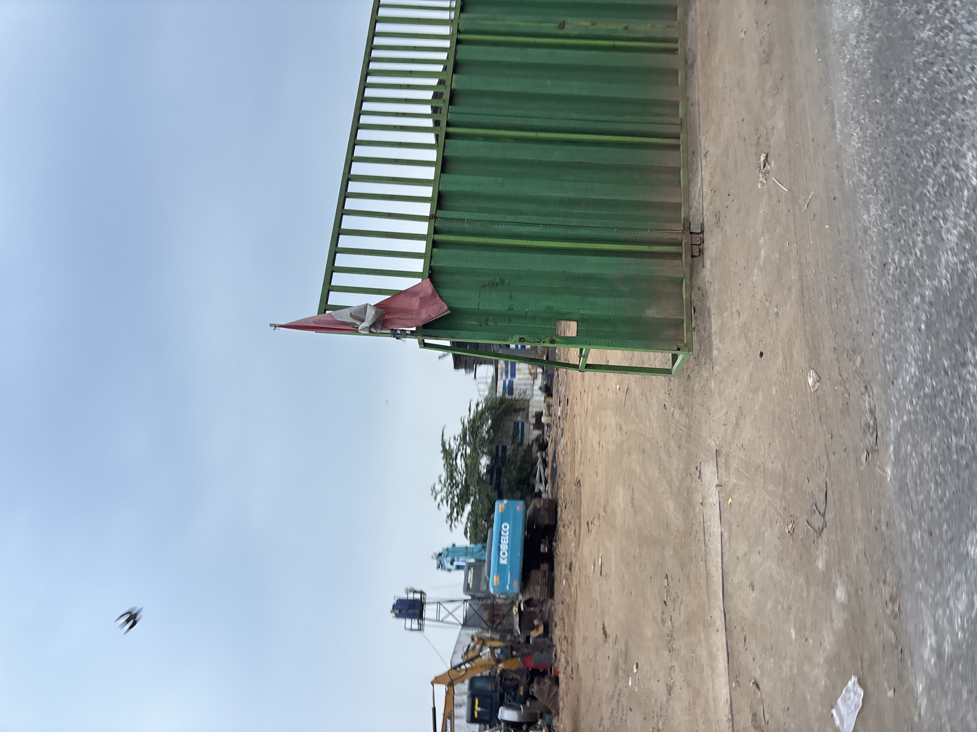 An exterior, ground-level, wide shot shows a construction or industrial site in Bekasi, Indonesia, under a cloudy sky. In the foreground, a large, green metal gate is partially open, revealing a dirt and gravel ground surface with some debris. A red and white flag is attached to the top of the gate. Beyond the gate, construction equipment is visible, including a blue Kobelco excavator and a yellow DANY excavator. A water tower and other structures are also present in the background. A bird is in flight in the upper left quadrant of the image.